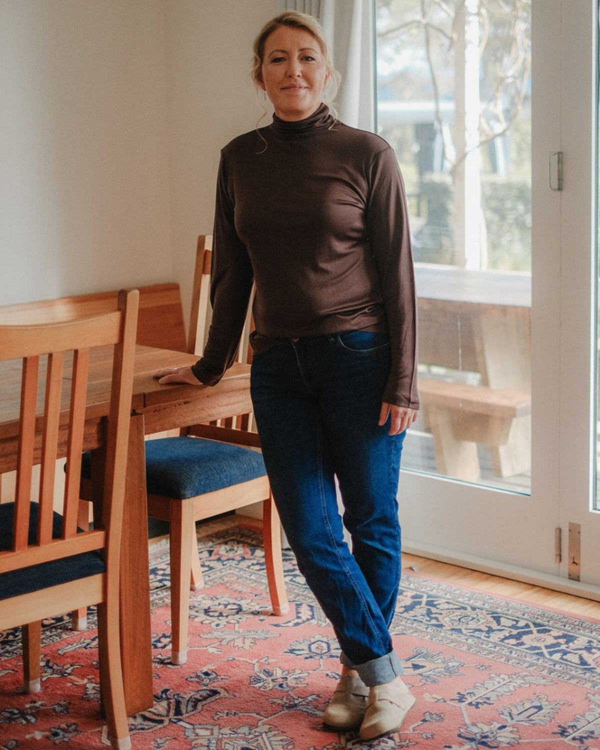 silk mahogany turtle neck Woman standing in a room with wooden furniture and a large window.
