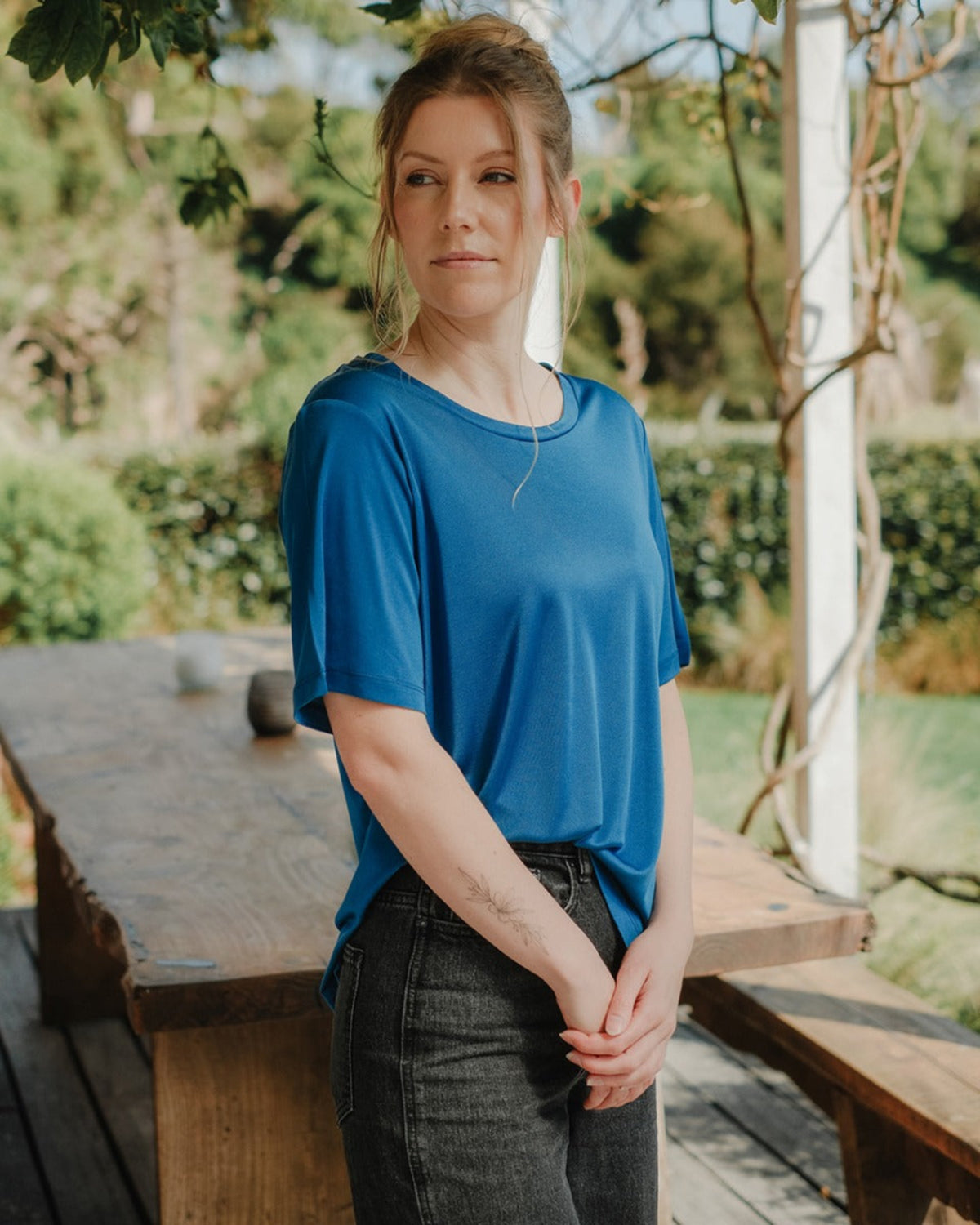 Woman in a silk blue shirt standing outdoors with greenery in the background