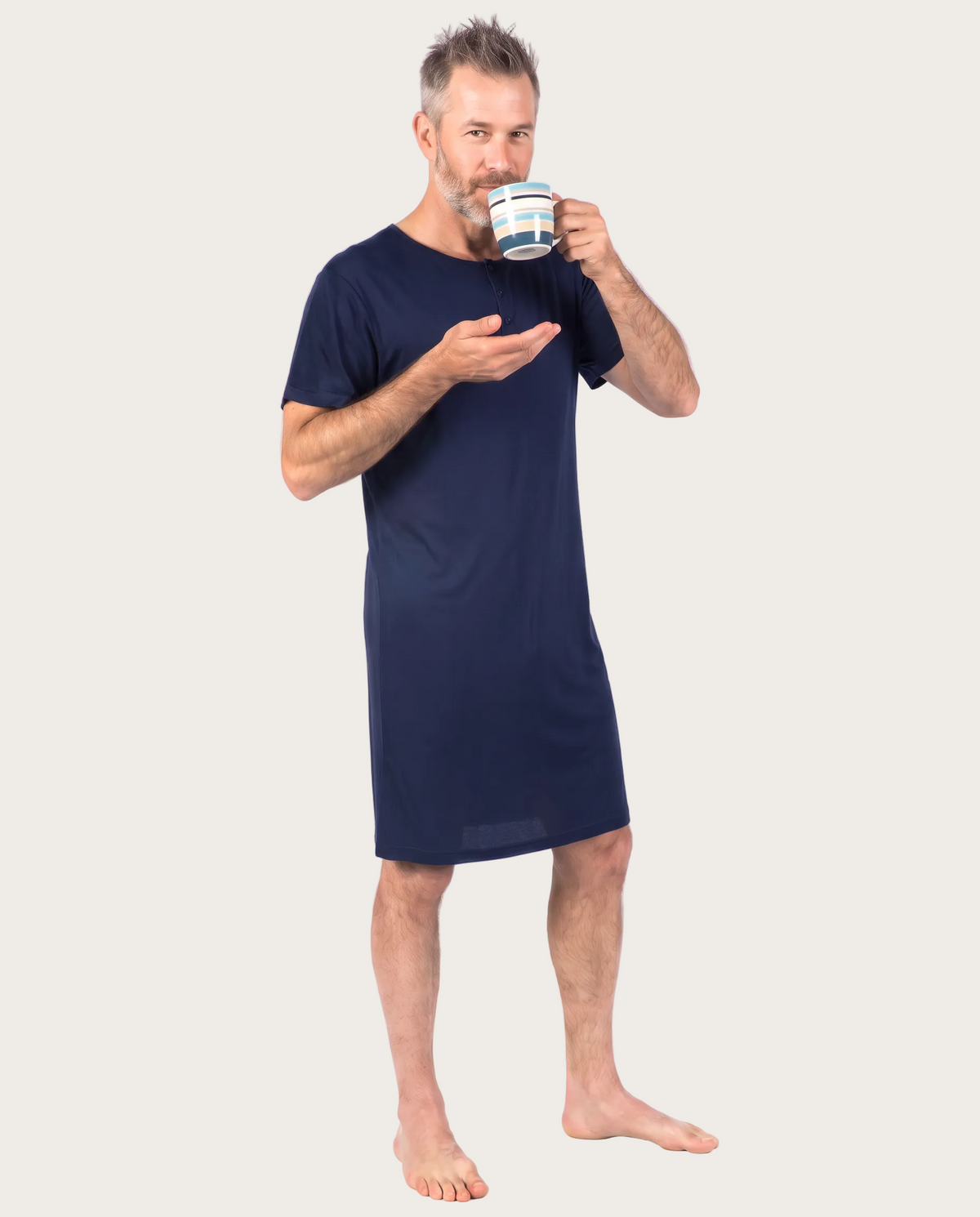 A man wearing a navy blue short-sleeve nightshirt or pajama dress, holding a striped mug, standing against a plain white background.