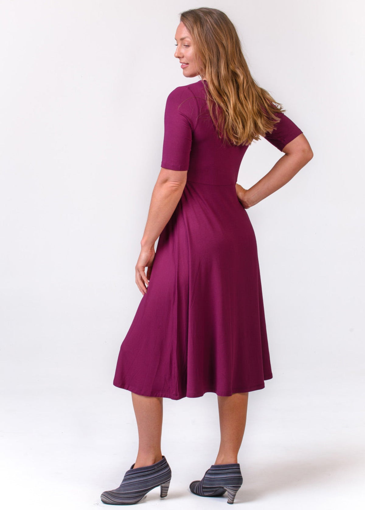 A woman modeling a short-sleeve, knee-length A-line dress in a deep berry purple, shown from a side-back angle against a plain white studio background.