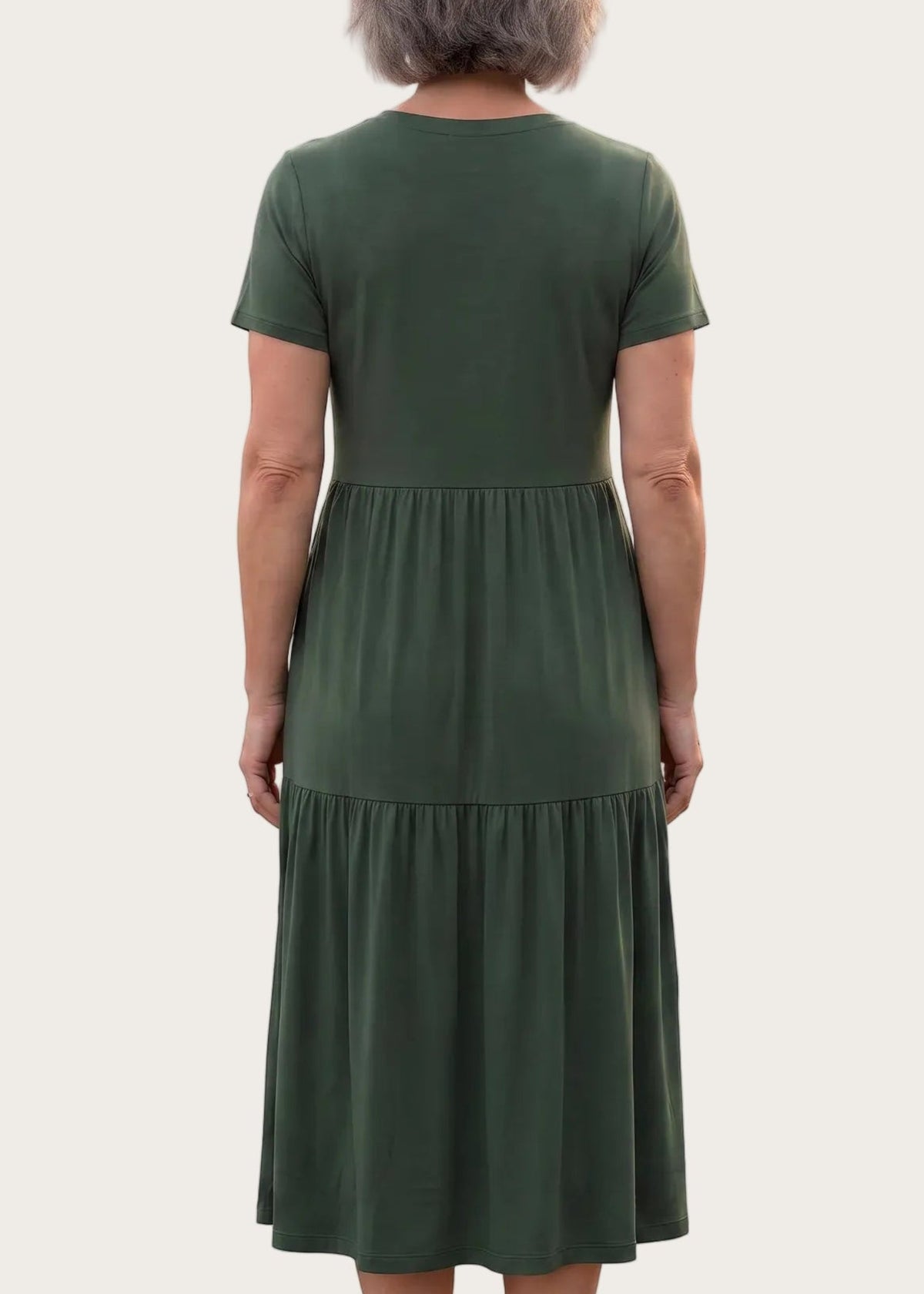 An older woman viewed from the back wearing a short-sleeve, tiered midi dress in dark olive green, paired with brown wedge sandals, against a plain white background.