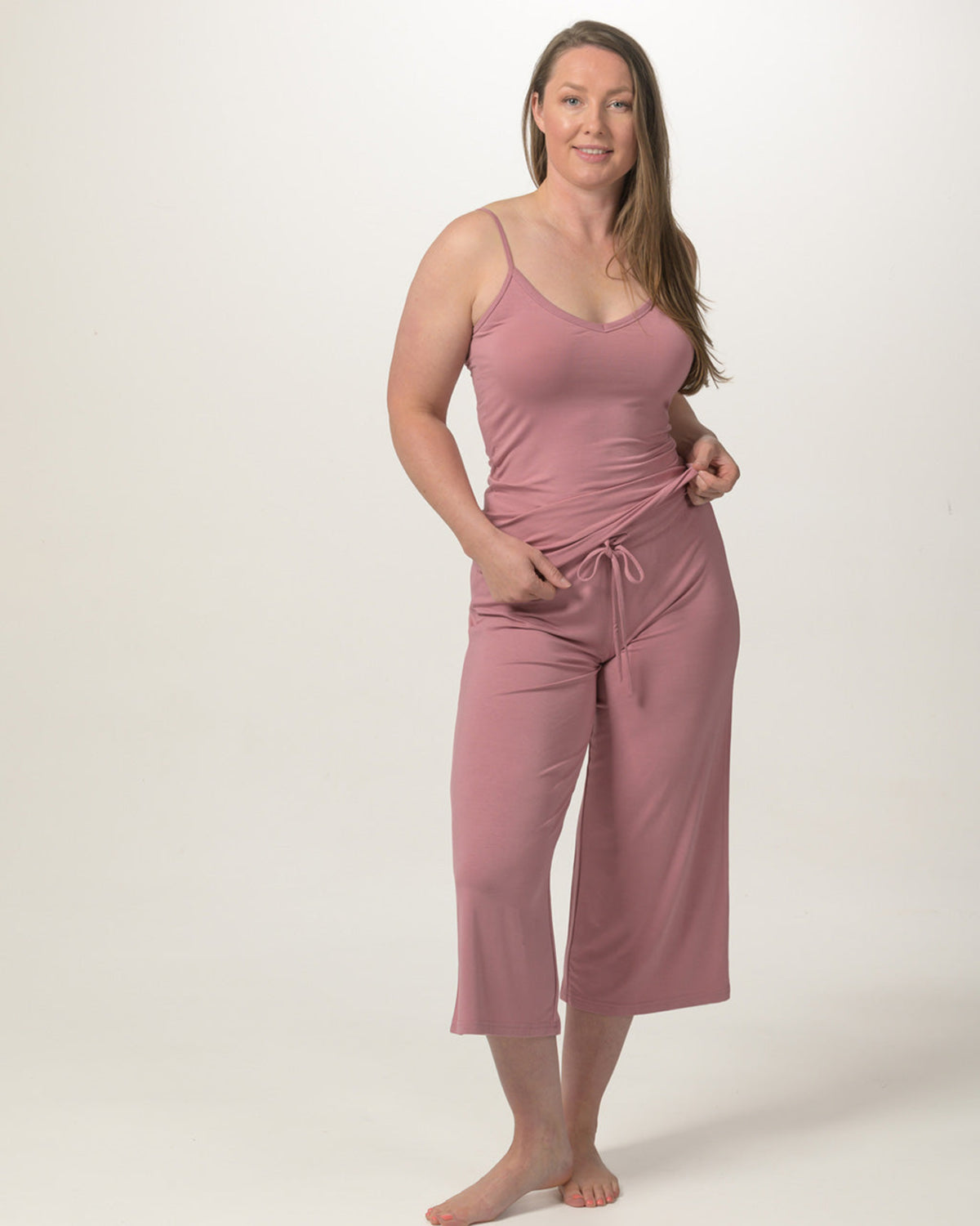 A woman modeling a dusty rose two-piece loungewear set consisting of a spaghetti-strap bamboo camisole top and matching cropped drawstring pants, shot against a plain light gray studio background.