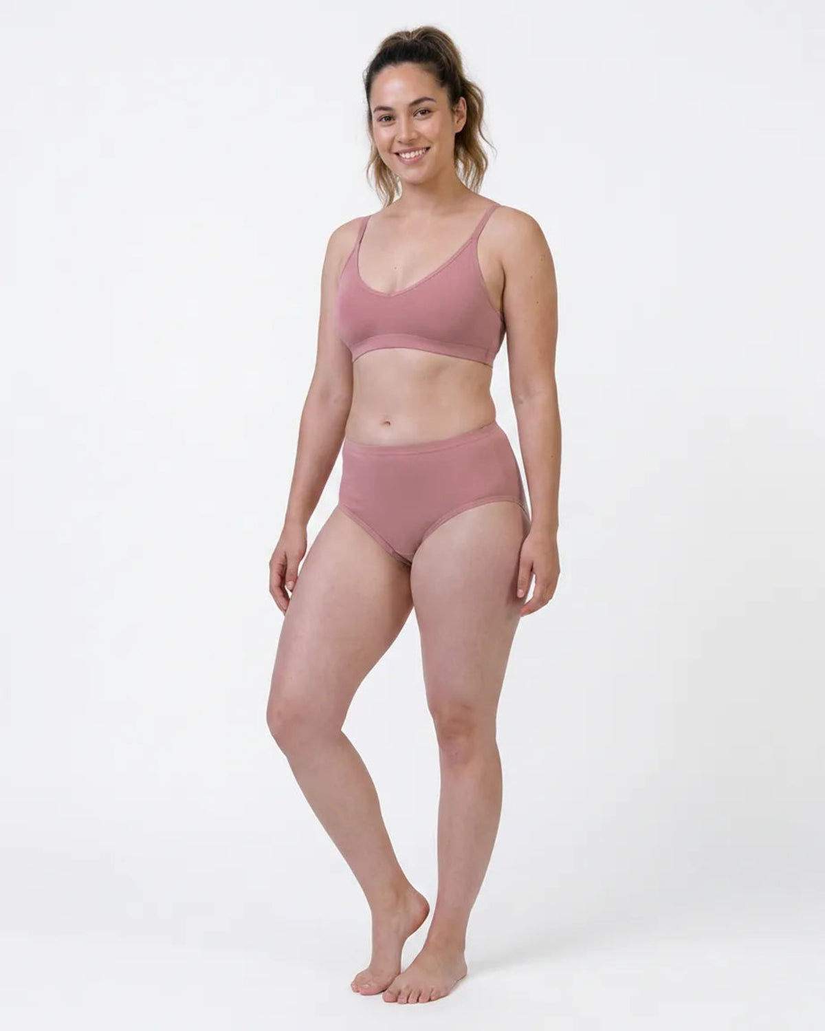 A woman modeling a dusty rose two-piece lingerie set consisting of a scoop-neck bralette and matching high-waisted briefs, photographed against a seamless white studio background.