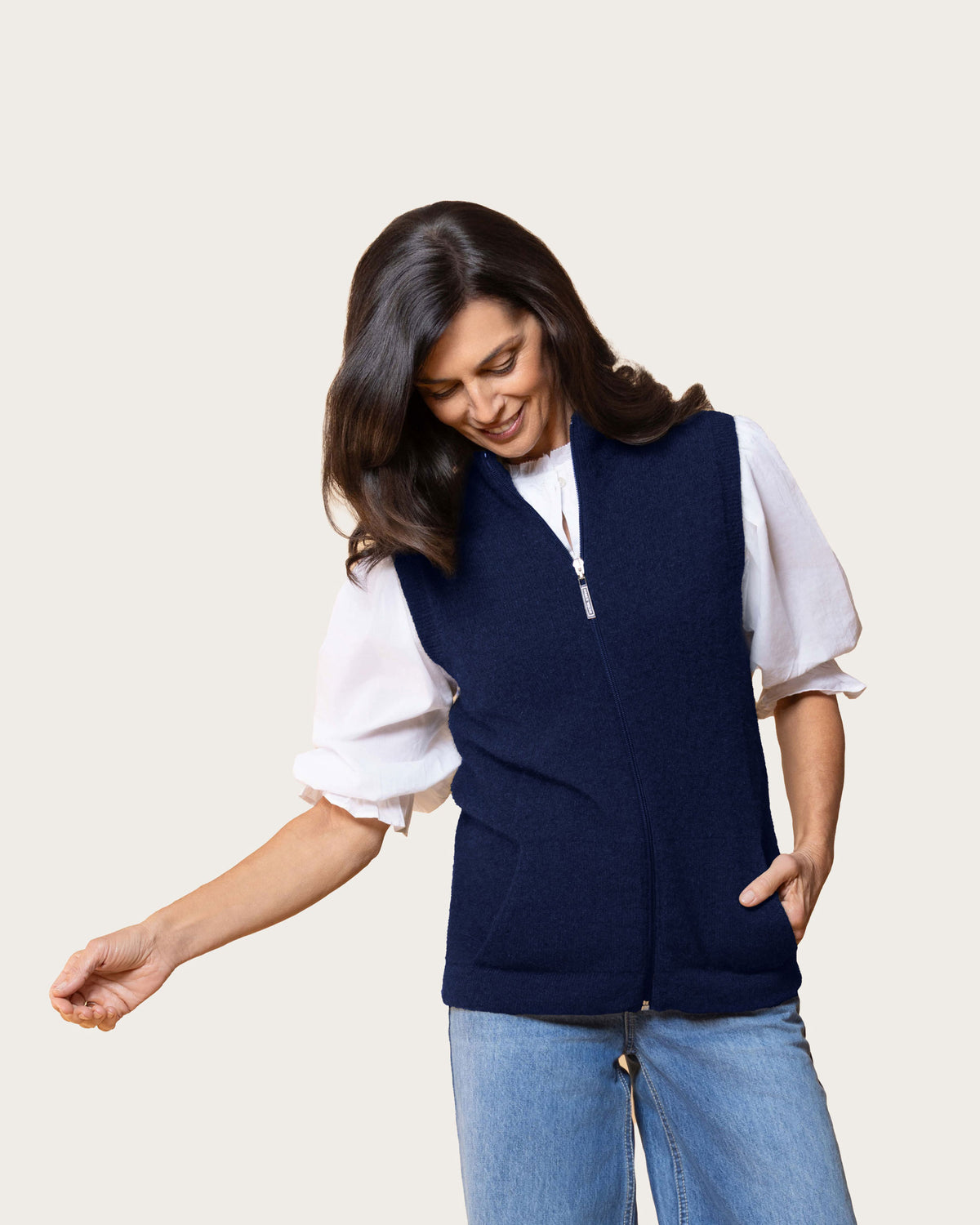 A woman wearing a navy blue sleeveless zip-front  possum merino vest over a white puff-sleeve blouse and light blue jeans, photographed against a plain light beige studio background.