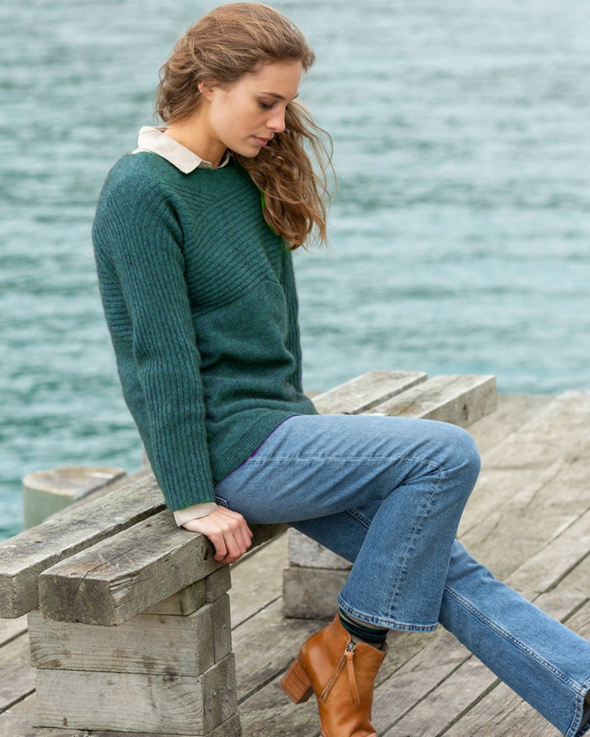 possum merino Woman sitting on a wooden bench by the water wearing a green sweater and blue jeans.