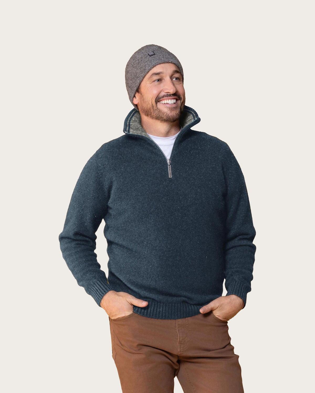A man wearing a denim blue possum merino quarter-zip knit sweater with a gray beanie and brown pants, standing against a solid light beige background.