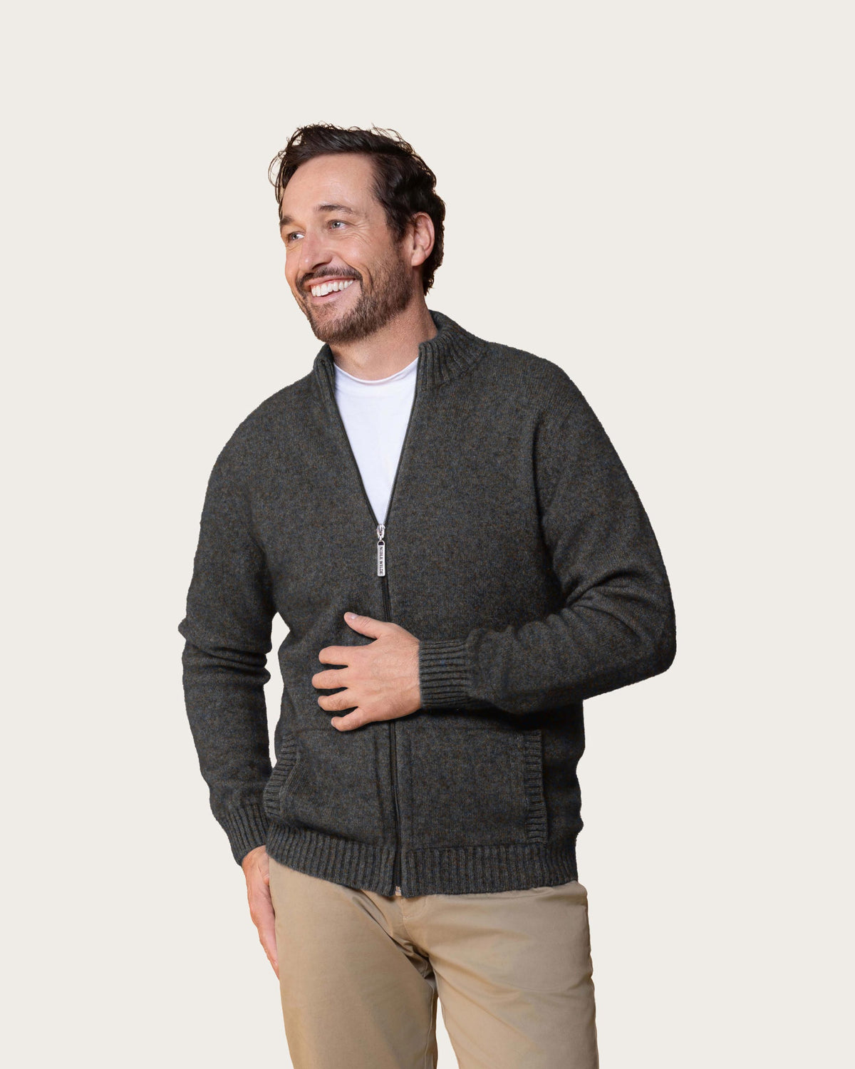 A smiling man wearing a dark melange gray zip-up possum merino knit sweater over a white t-shirt and khaki pants, photographed against a solid light beige background.