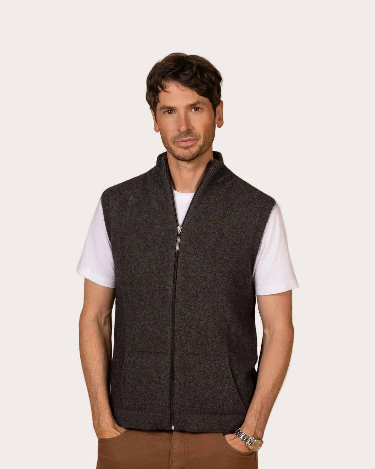possum merino A man wearing a dark charcoal gray zip-up knit vest over a white short-sleeve t-shirt and brown pants, standing against a solid off-white background.