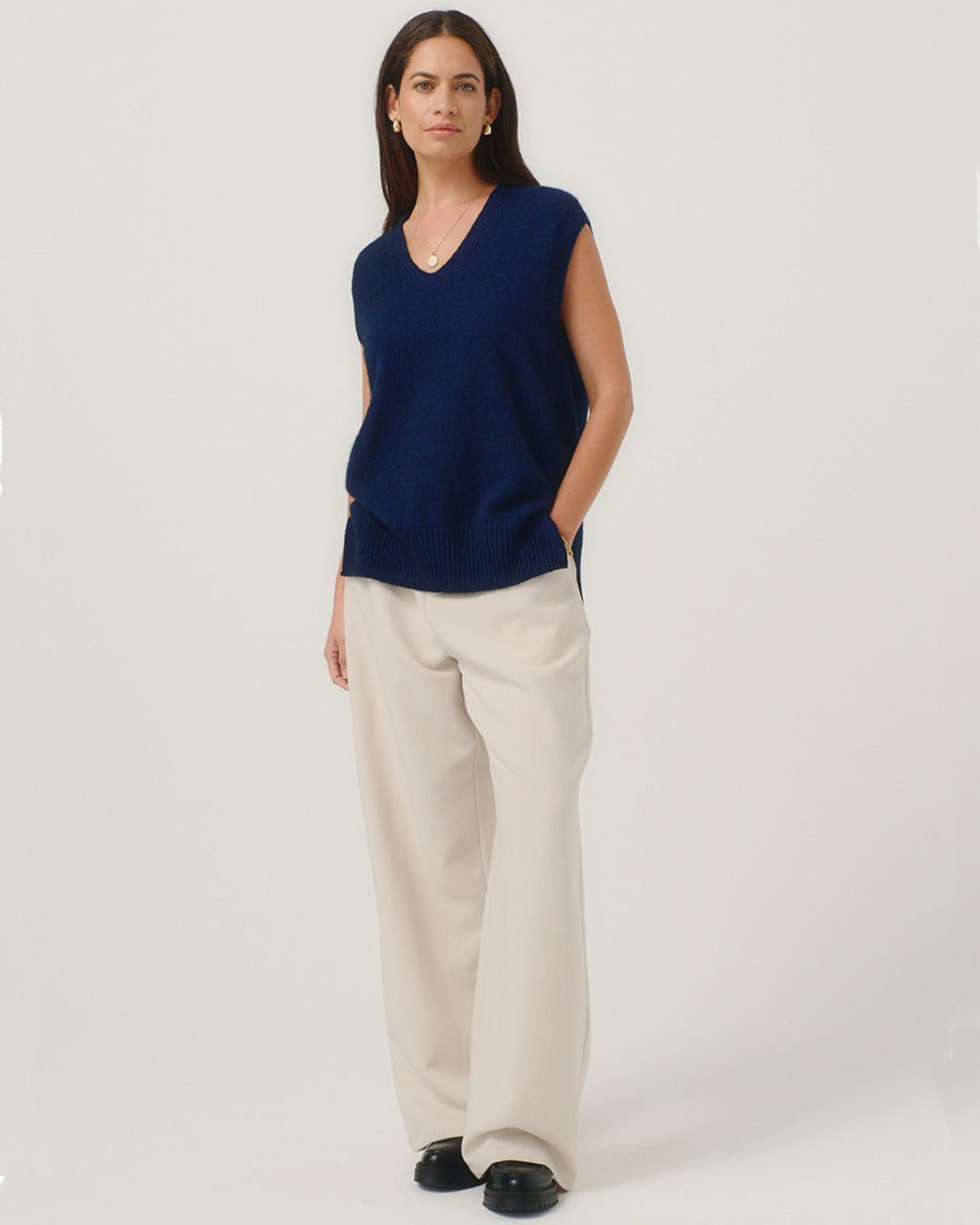 Possum Merino Woman wearing a blue sleeveless top and beige pants on a white background