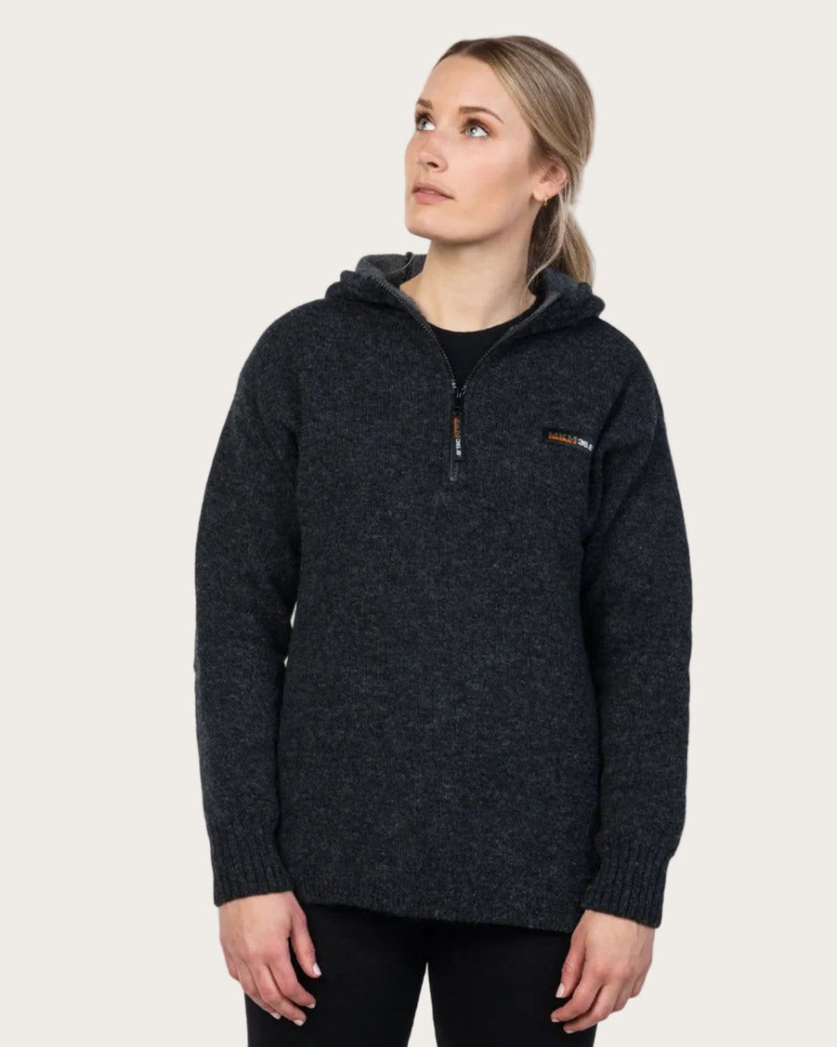 Wool A woman wearing a dark charcoal heathered pullover hoodie with a half-zip chest and a small orange and white logo on the left chest, photographed against a solid light beige background.