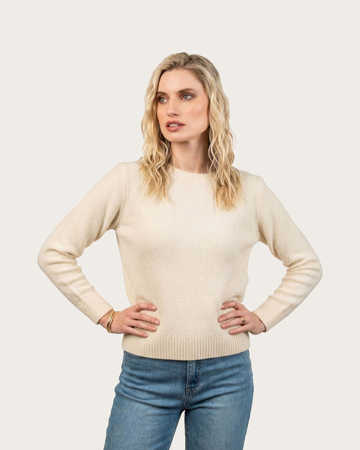 A blonde woman modeling a cream-colored Possum Merino knit sweater with blue jeans, standing against a solid light beige background.