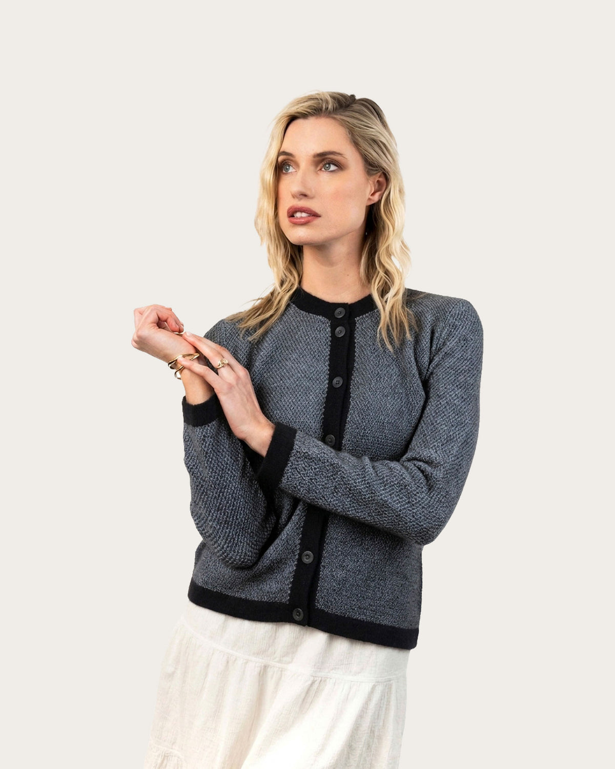 A blonde woman wearing a textured black and white, possum merino cardigan with black trim and buttons, paired with a white skirt, posing against a solid off-white studio background.