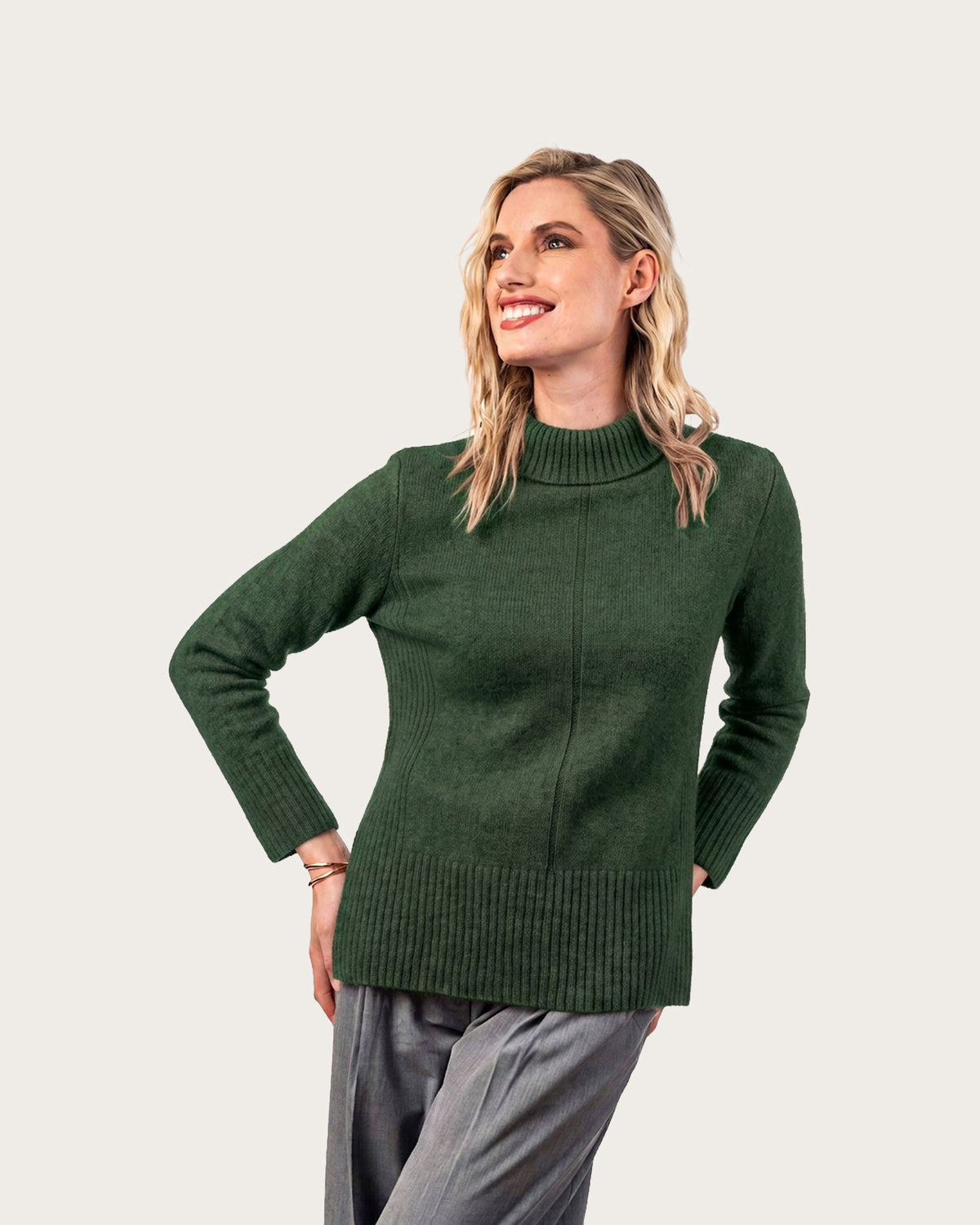 A woman modeling a dark green ribbed mock neck possum merino sweater with long sleeves, paired with gray trousers, against a solid light beige background.