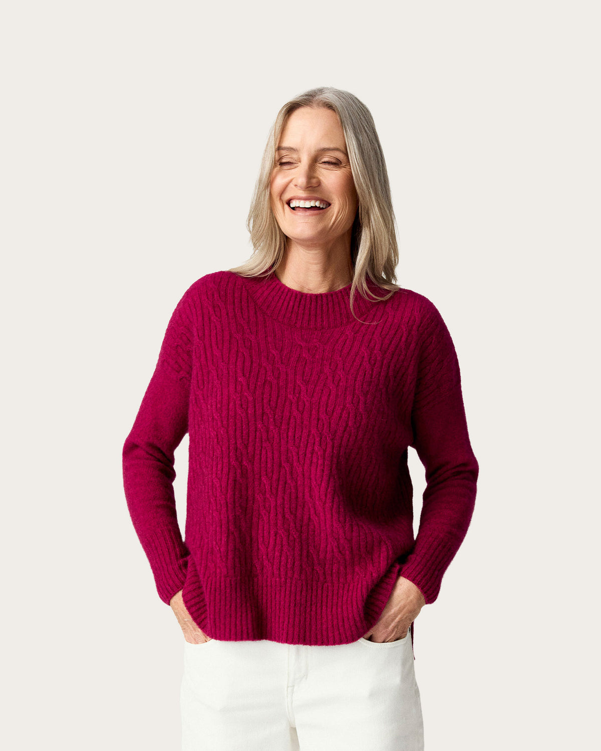 A smiling woman with blonde hair wearing a chunky cable-knit crewneck possum merino sweater in deep magenta and white pants, standing against a solid light beige background.