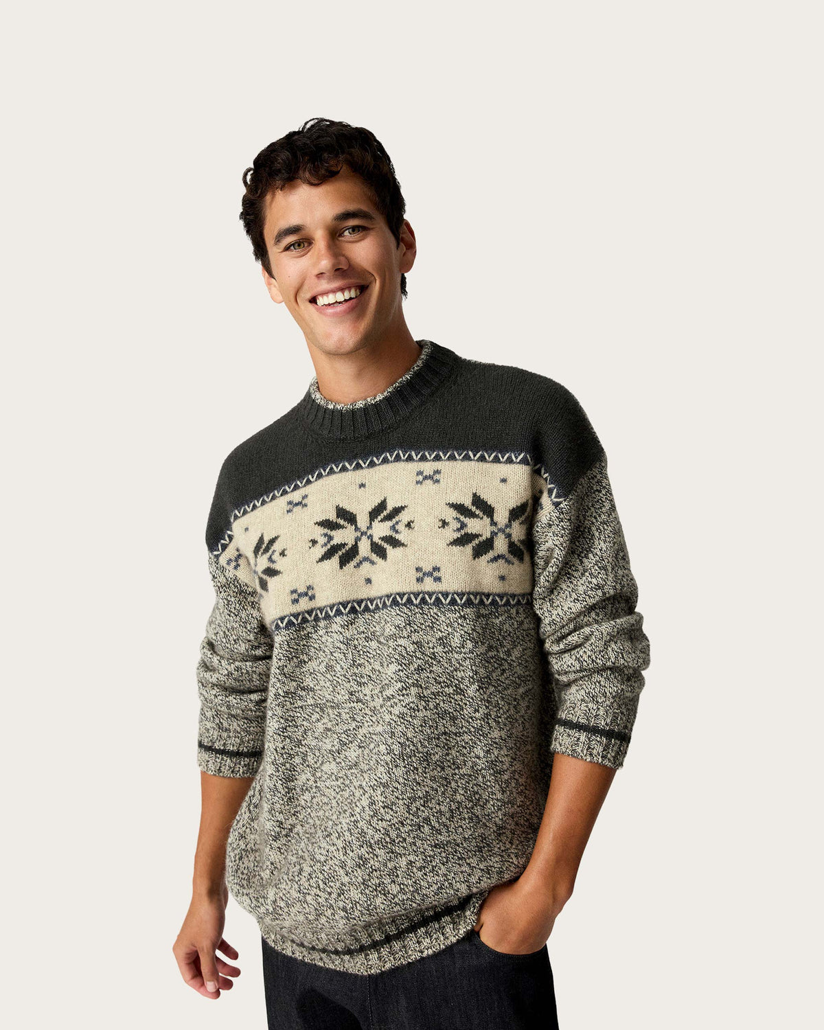 A man wearing a knit crewneck possum merino sweater with a dark gray yoke, a cream horizontal band with black snowflake patterns, and a heather gray lower body, posed against a solid light gray background.