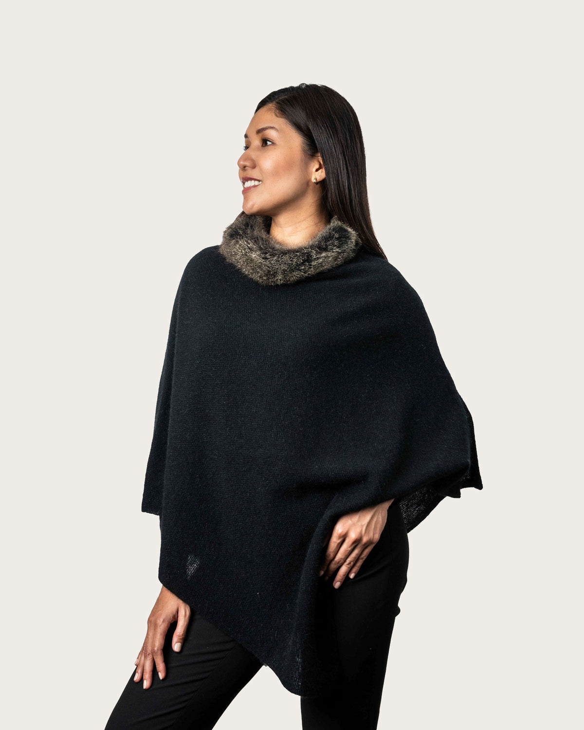 A woman wearing a black knit poncho with a gray possum fur collar, posed against a solid light beige background.