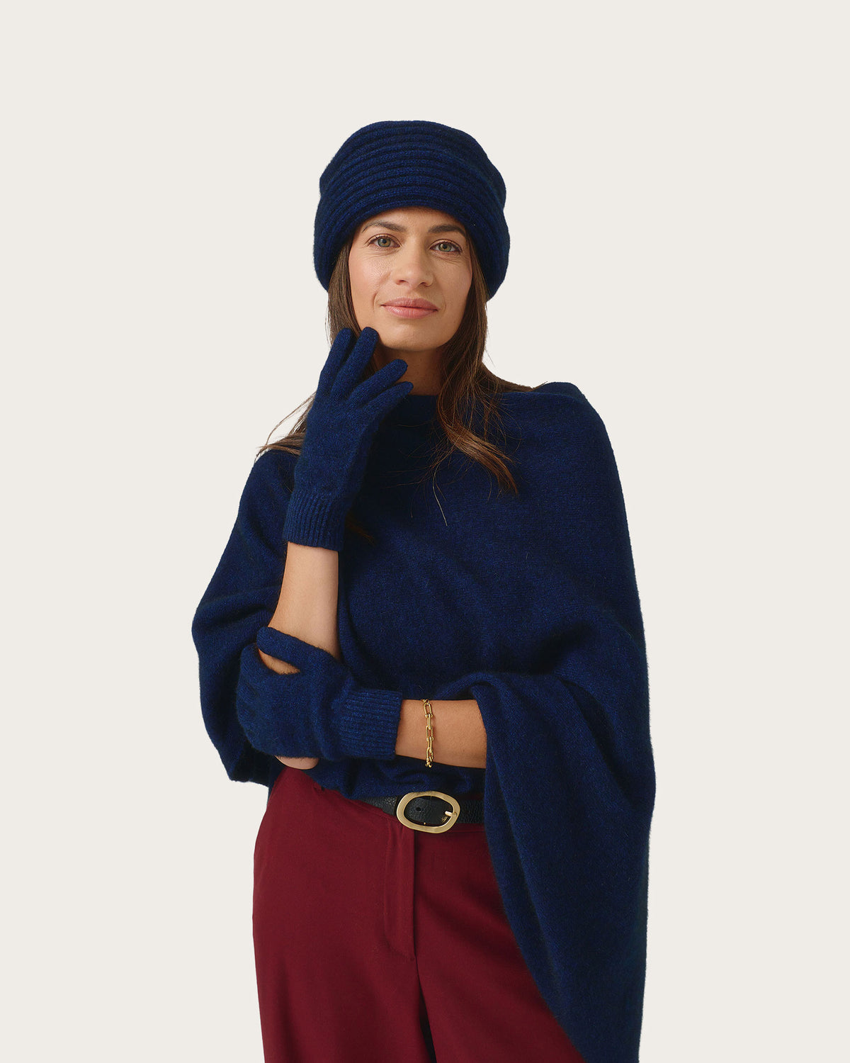 A woman wearing a navy blue knit beanie, matching knit fingerless gloves, and a navy blue poncho-style sweater over burgundy trousers, photographed against a plain light gray studio background.