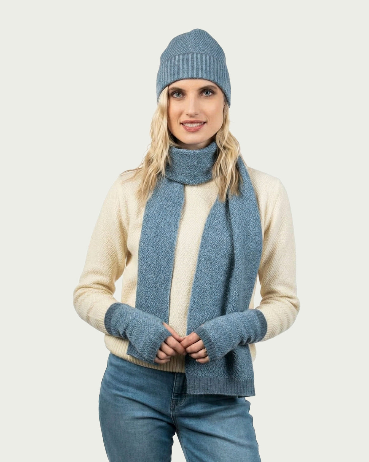 A woman modeling a matching knit winter set consisting of a blue beanie, a blue possum merino scarf, blue fingerless gloves, and a cream-colored sweater, set against a solid light gray background.