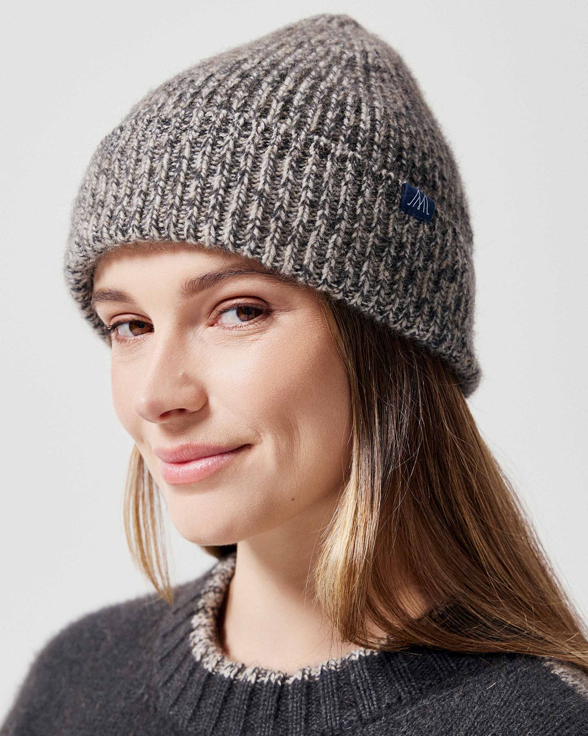 A woman wearing a gray and white marled ribbed possum merino knit beanie hat with a small blue embroidered logo patch on the side, photographed against a light gray studio background.