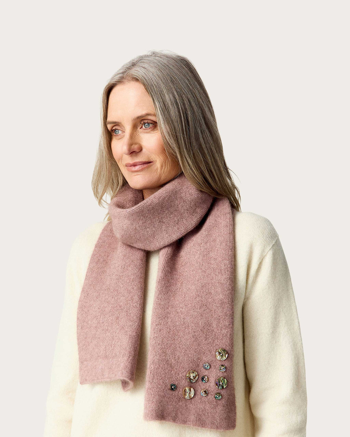 A woman with gray hair wearing a dusty rose possum merino knit scarf with decorative metallic buttons, paired with a cream sweater, against a light gray background.
