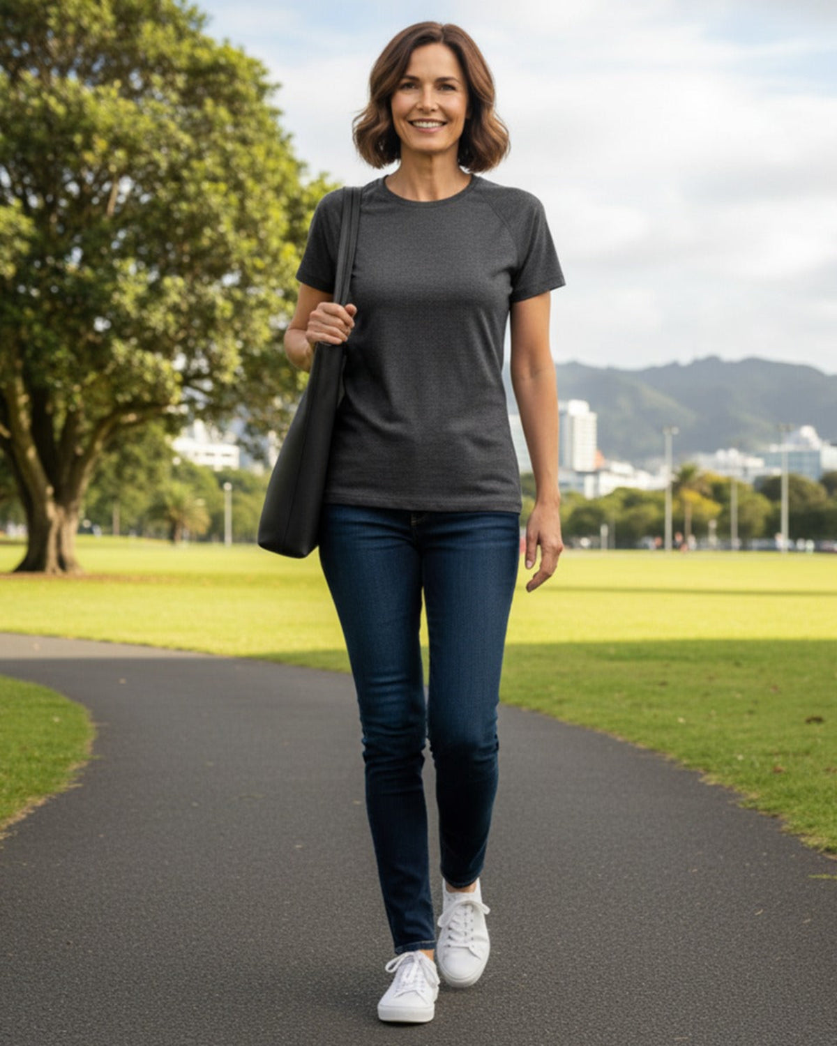 Woman walking on a path in a park wearing a merino dark gray t-shirt, blue jeans, and white sneakers.