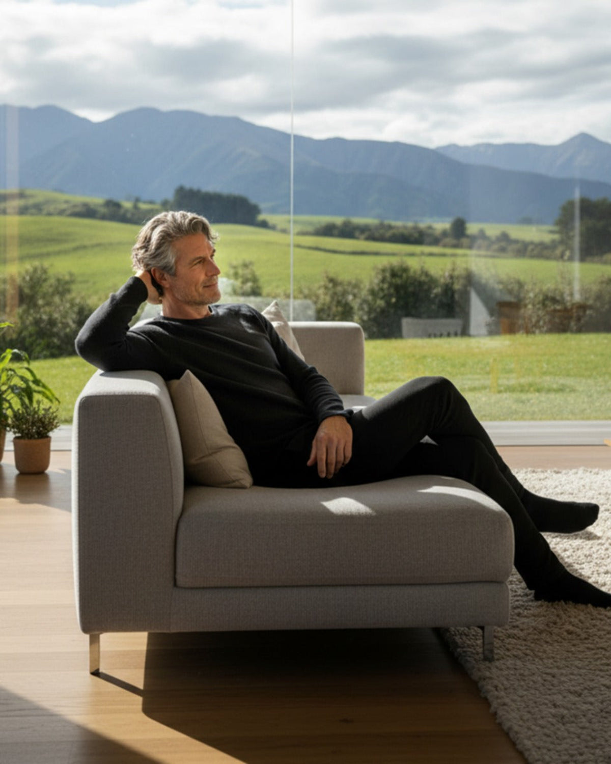 Acrux Superfine Merino Long Sleeve T-Shirt Man sitting on a couch in a modern living room with a scenic view of mountains and fields.