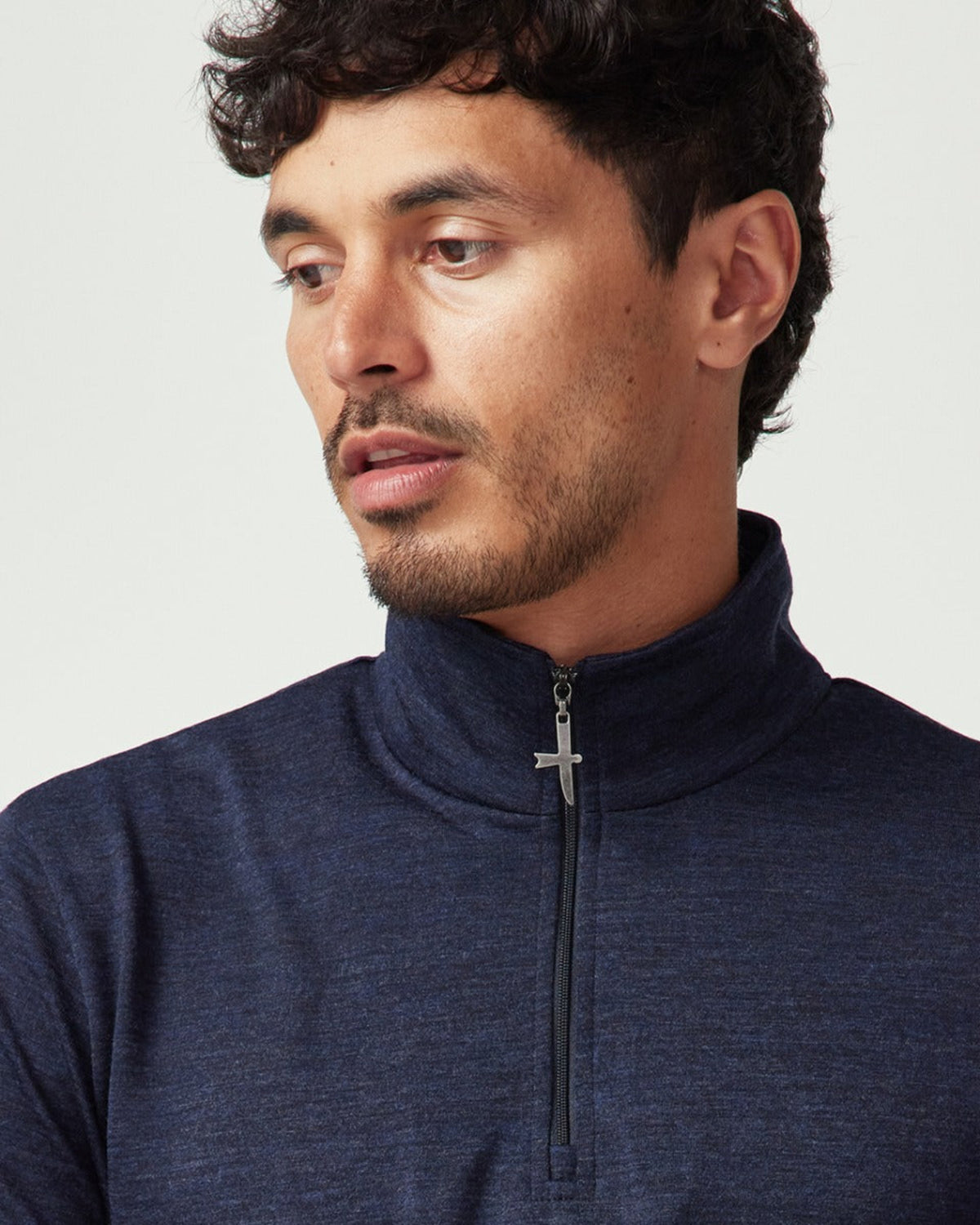 Merino Man wearing a navy blue zip-up sweatshirt with a cross pendant on a light gray background