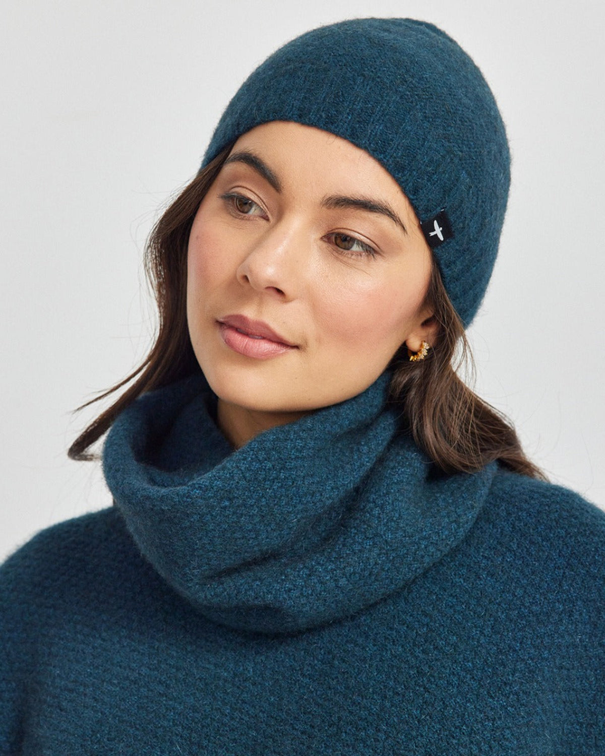 Possum Merino Woman wearing a teal knit beanie and scarf on a light gray background