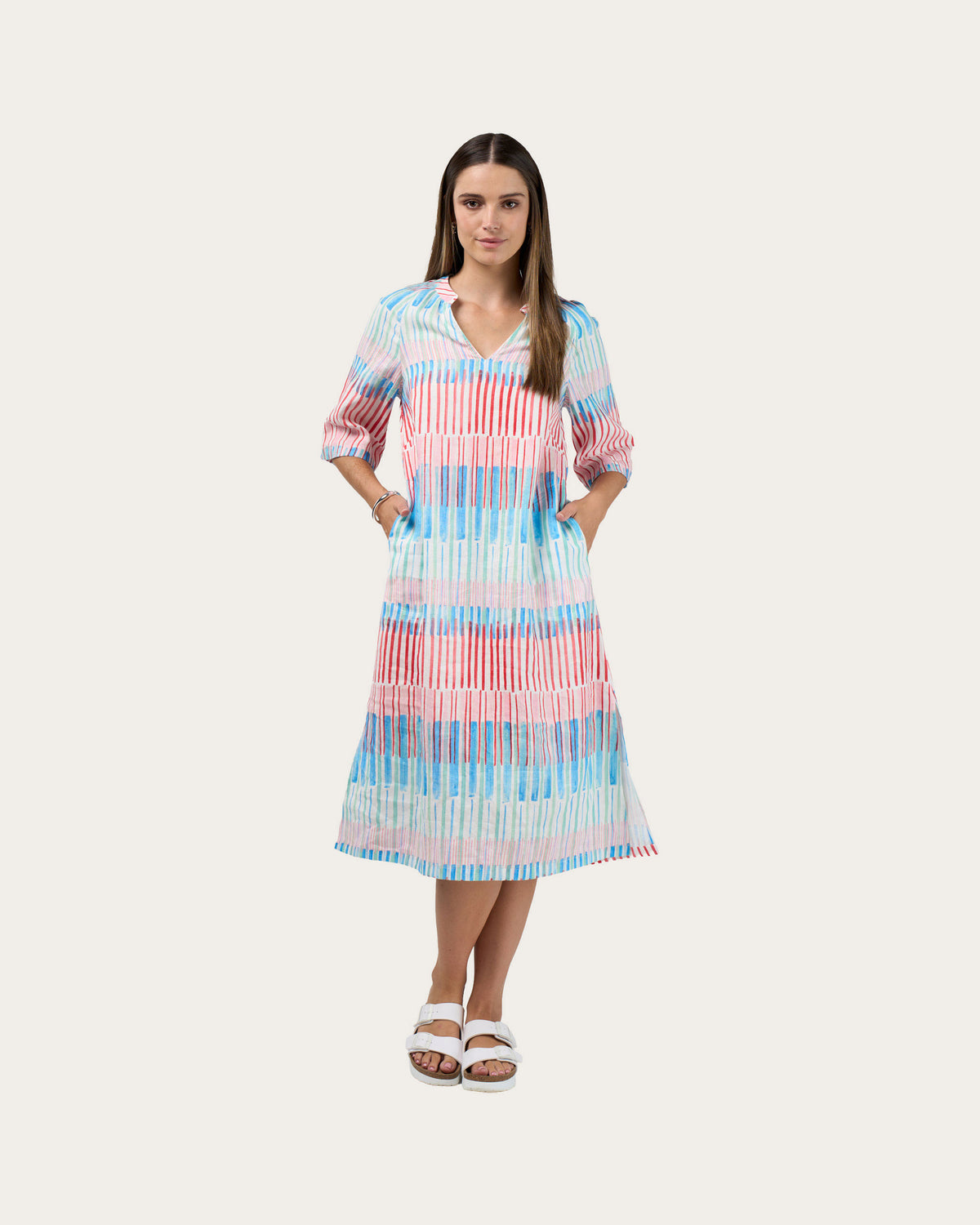 A woman modeling a linen midi dress with a vertical striped pattern in pink, blue, and white, paired with white slide sandals, set against a solid light beige background.