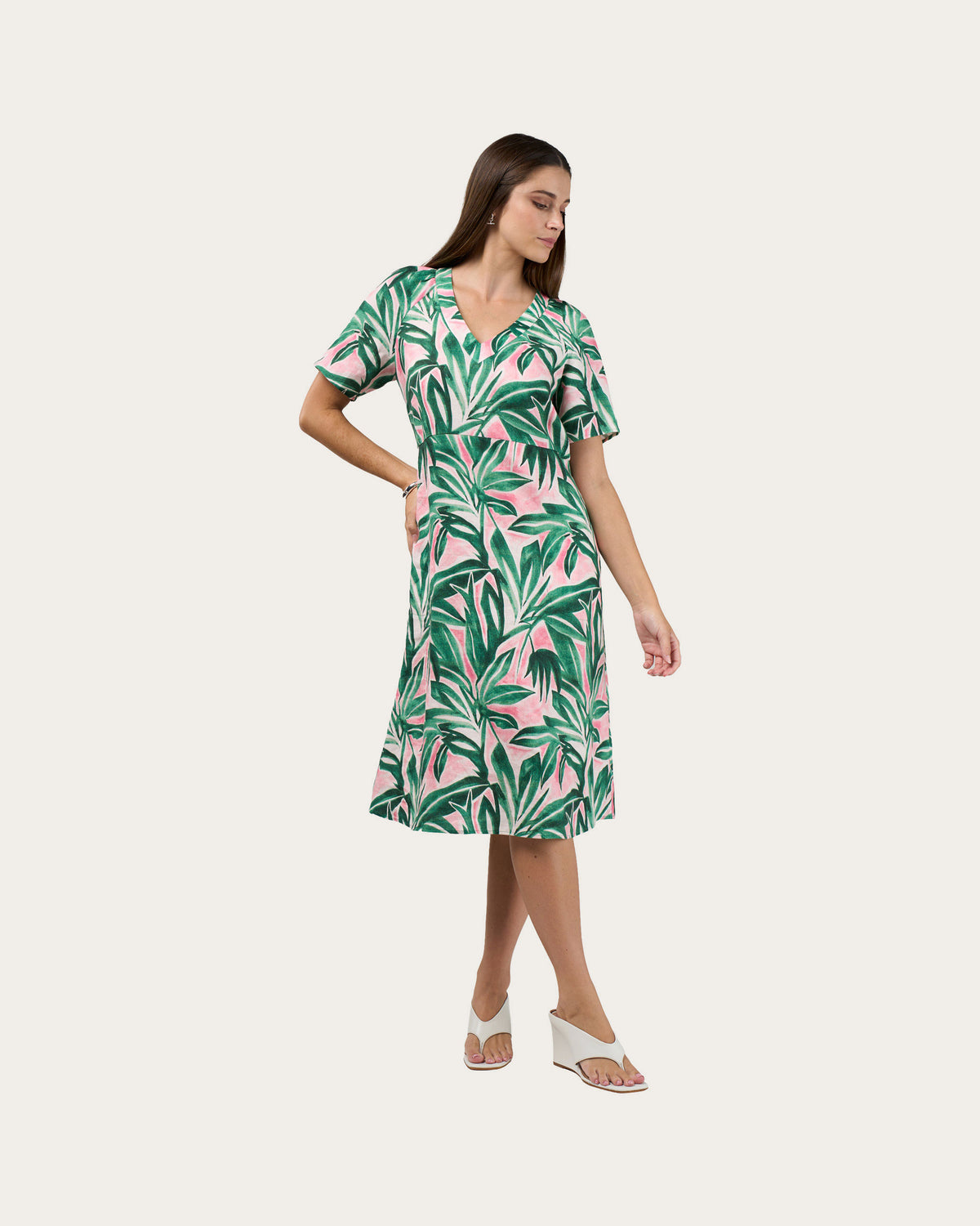 A woman modeling a short-sleeve linen midi dress with a green and pink tropical leaf pattern, paired with white wedge sandals, against a solid light beige background.