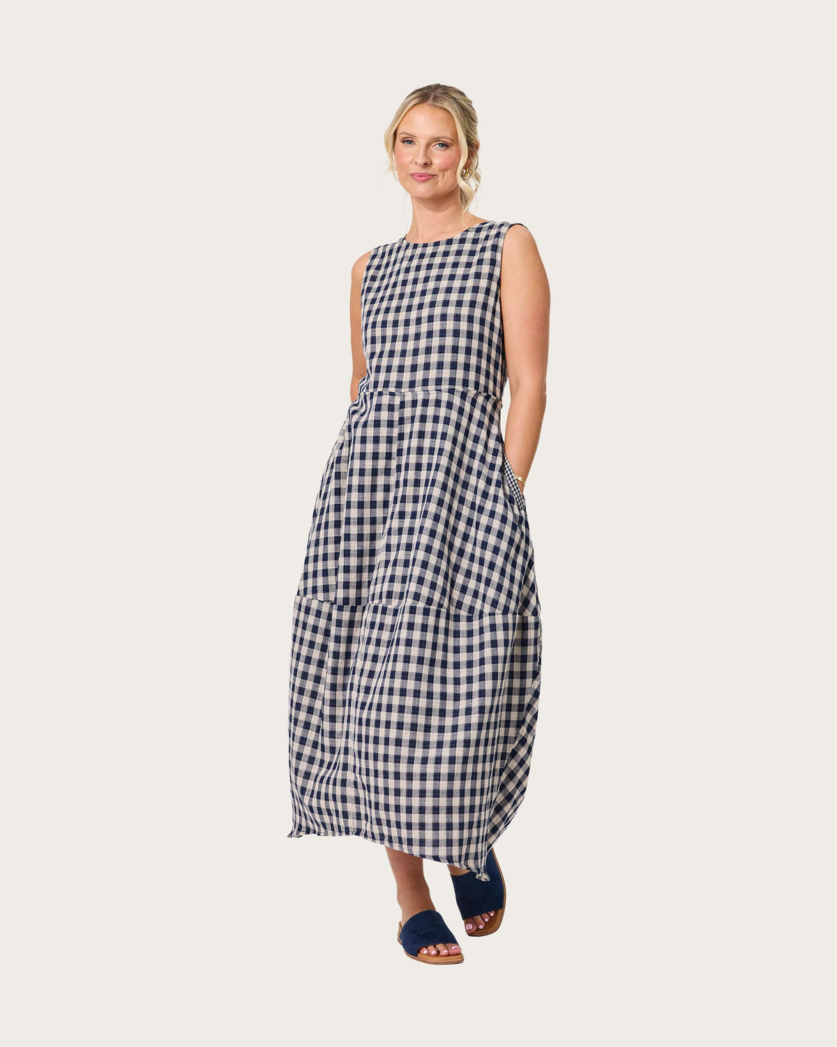 A woman modeling a sleeveless, tiered maxi dress with a navy and beige gingham check pattern, by Lemon Tree Design. Paired with dark blue slide sandals, against a solid light beige background.