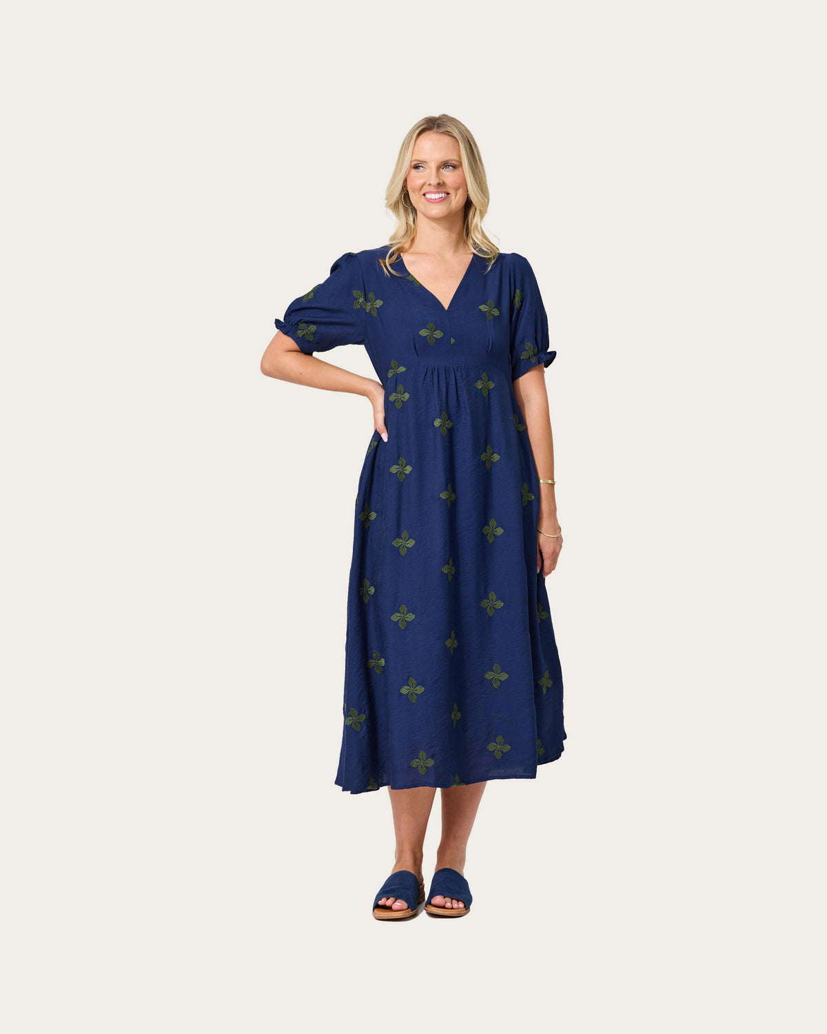A woman modeling a navy blue midi dress with a green fleur-de-lis pattern by Lemon Tree
, featuring puff sleeves and a V-neckline, set against a solid light gray background.