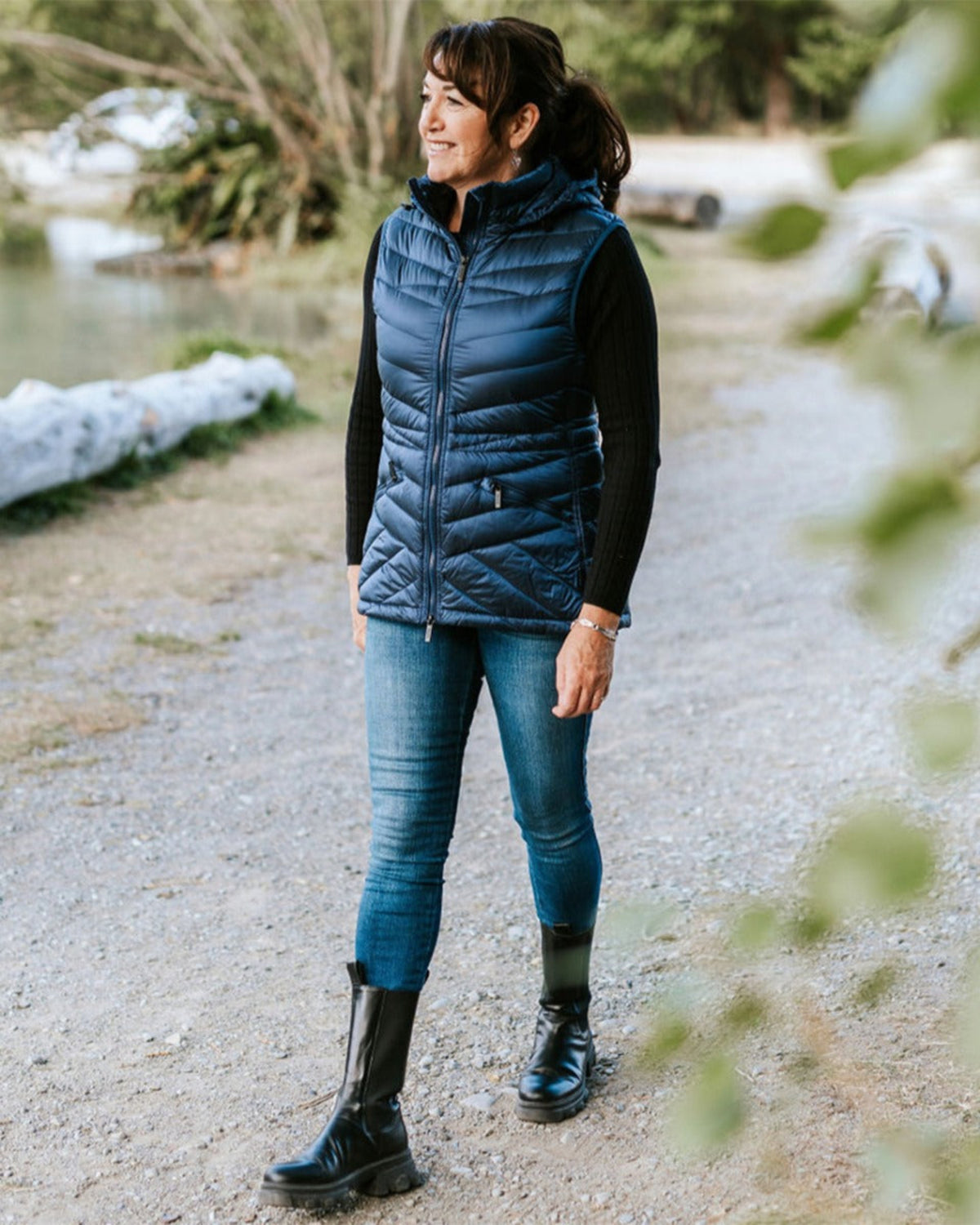 Woman wearing a blue puffer vest and jeans standing on a gravel path with a natural background.