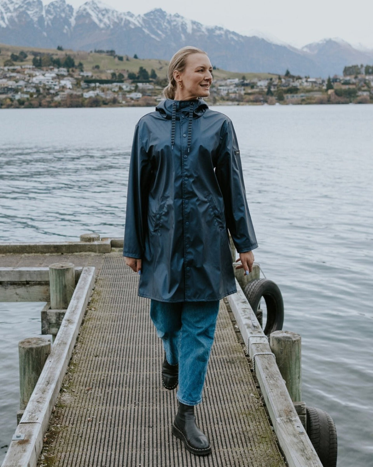 Person wearing a raincoat walking on a wooden dock by a lake with mountains in the background