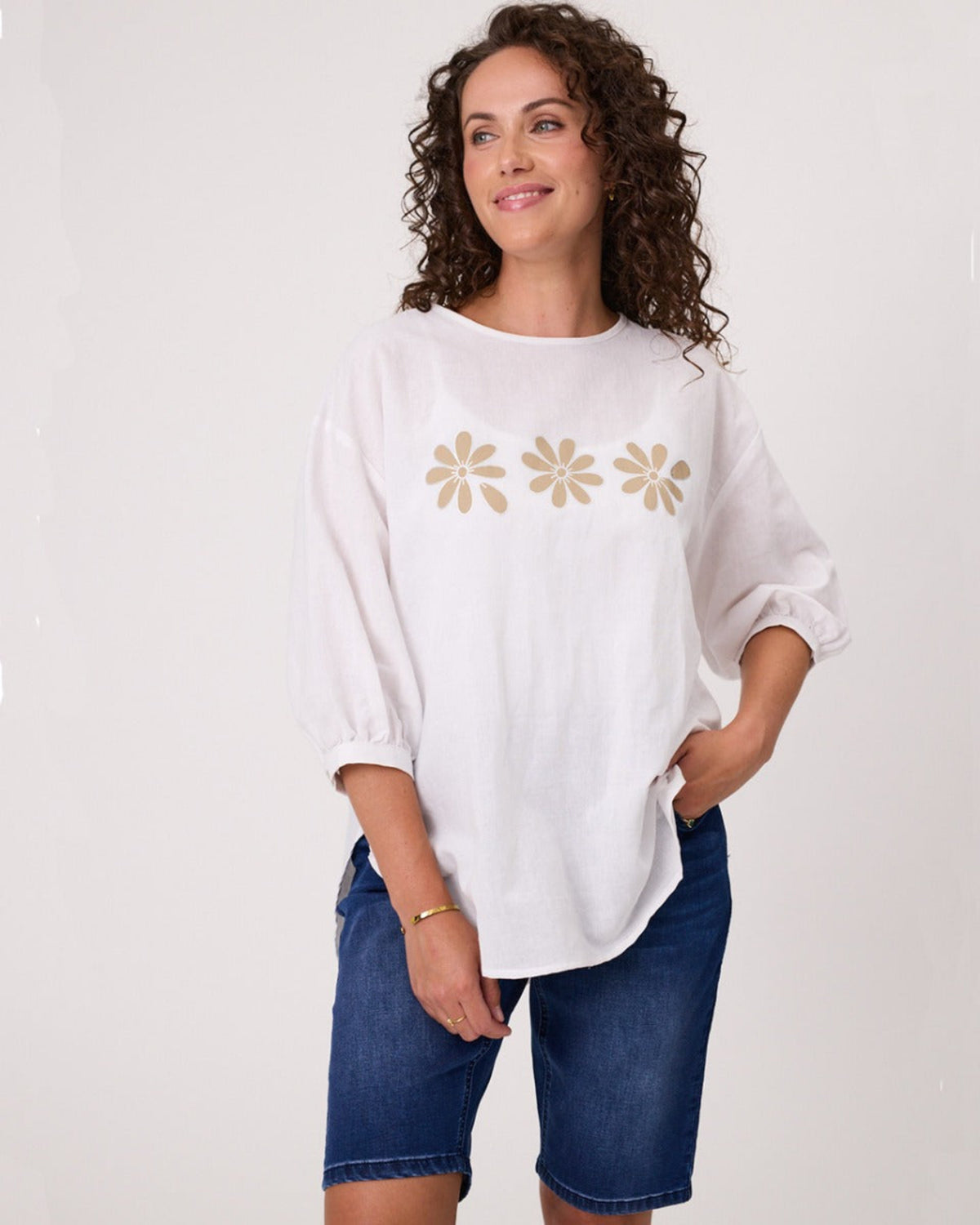 Woman wearing a white oversized shirt with floral designs and denim shorts on a plain background