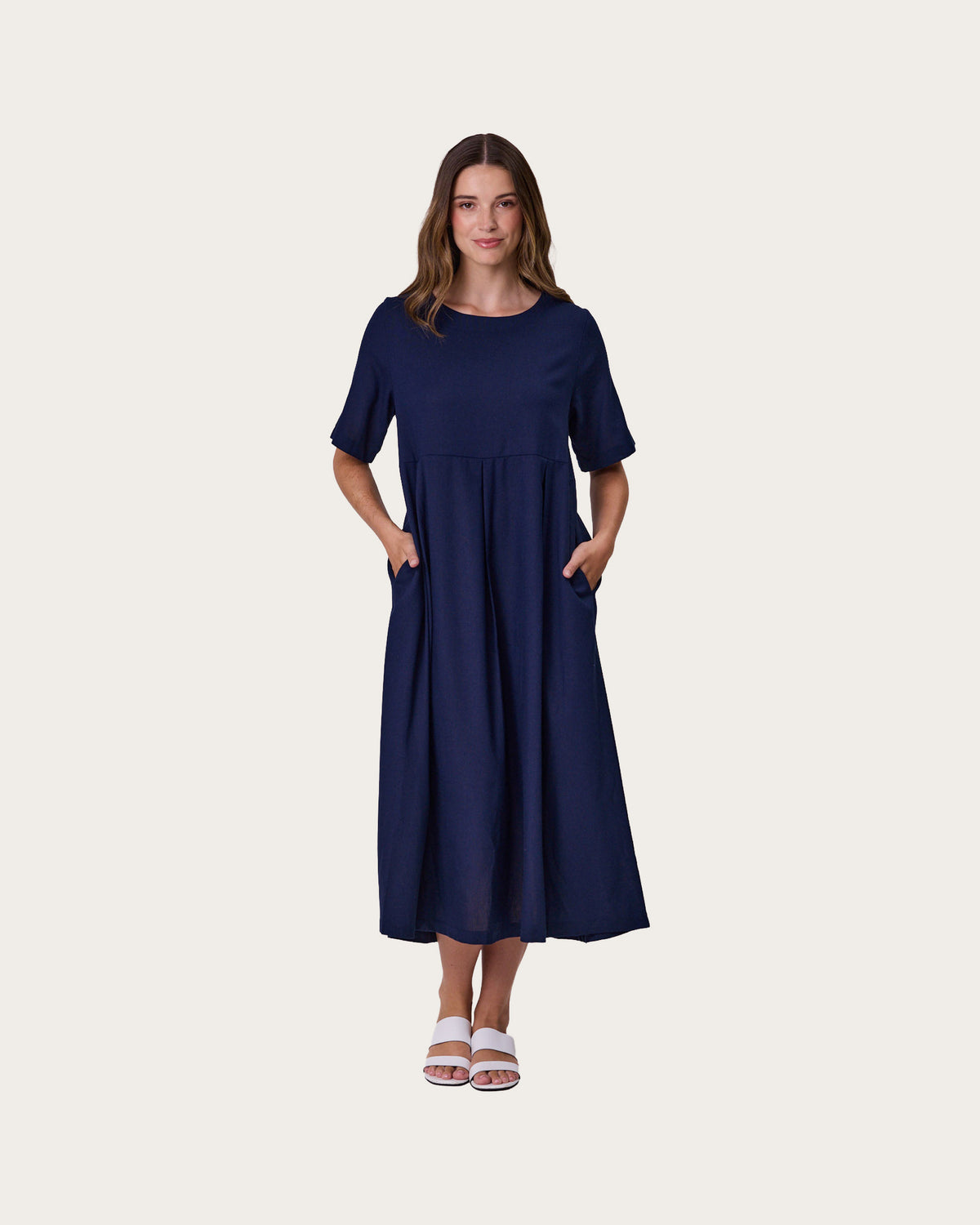A woman modeling a navy blue, linen blend, midi dress with short sleeves, a round neckline, and side pockets, standing against a solid off-white background.