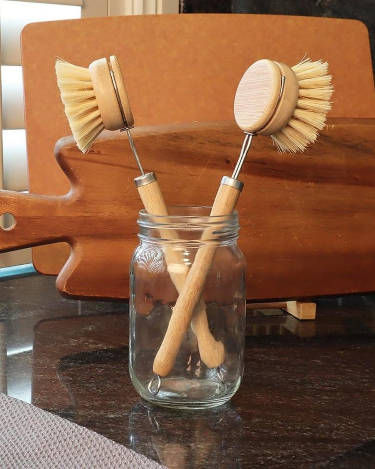 Three wooden dish brushes in a glass jar on a kitchen counter.