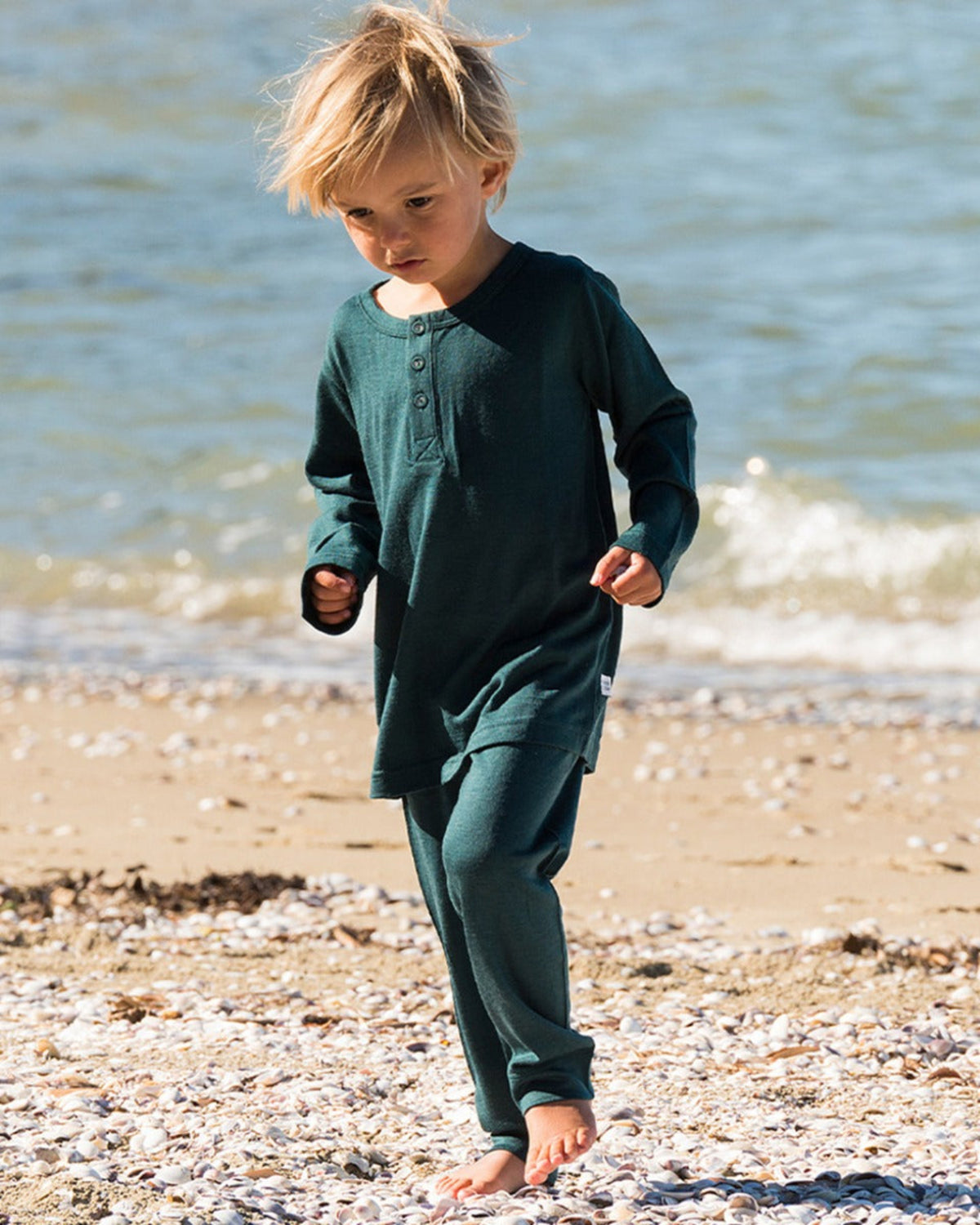 Child in merino green cuffed pants walking on a pebbly beach with water in the background