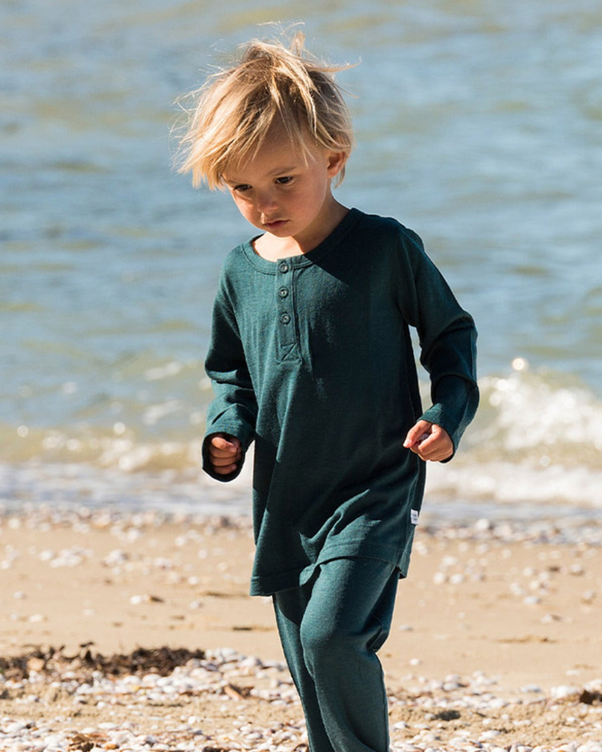 Merino Child in a green outfit standing on a beach with water in the background