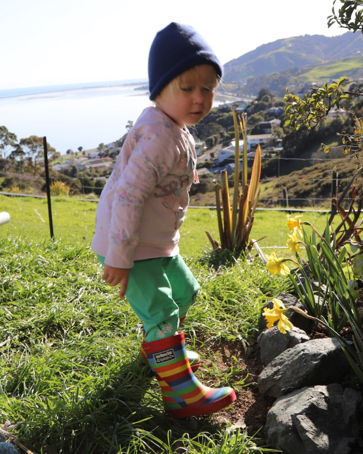 possum merino Child in colorful rain boots and a blue hat standing in a garden with flowers and rocks.