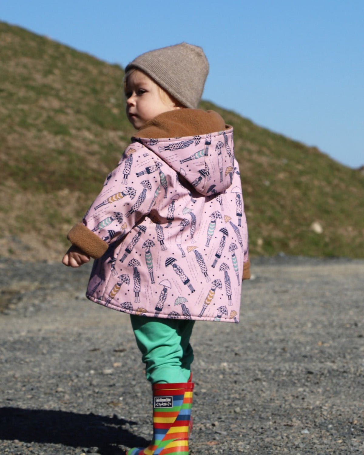 possum merino Child wearing a pink coat with a pattern, green pants, and rainbow boots outdoors.