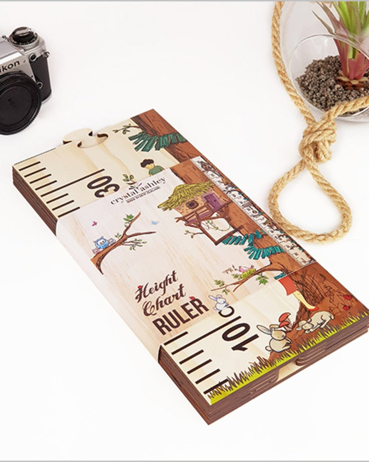 Wooden height chart ruler with cartoon designs on a white surface with a camera and plant pot in the background.