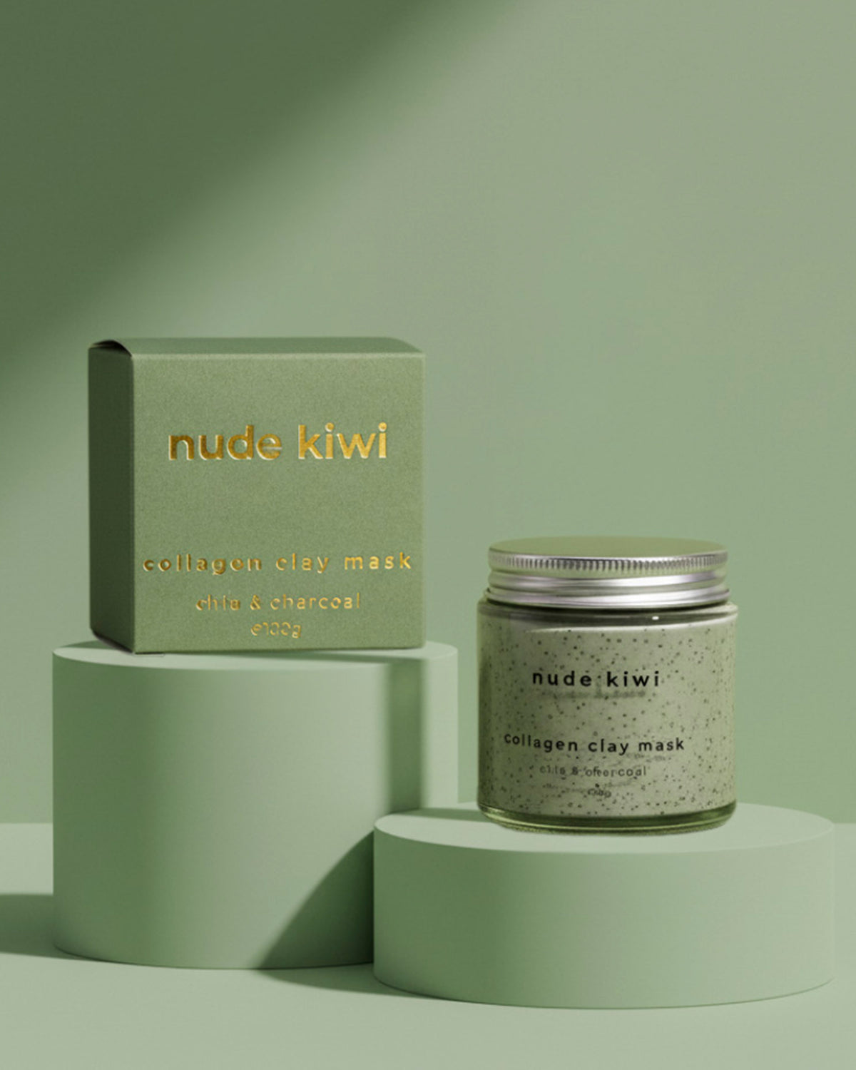 A jar of nude kiwi collagen clay mask with a silver lid sits next to its matching sage green box on minimalist green pedestal risers against a solid sage green background.