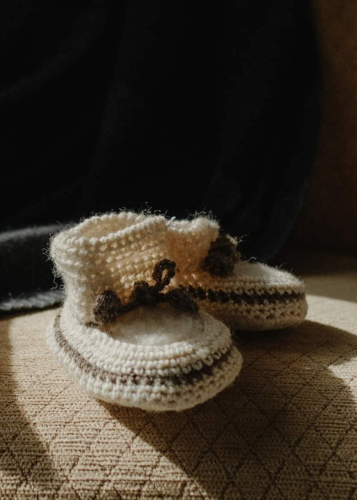 A pair of cream-colored crocheted baby booties with brown bows, resting on the seat of a beige textured armchair with a dark blanket draped in the background.