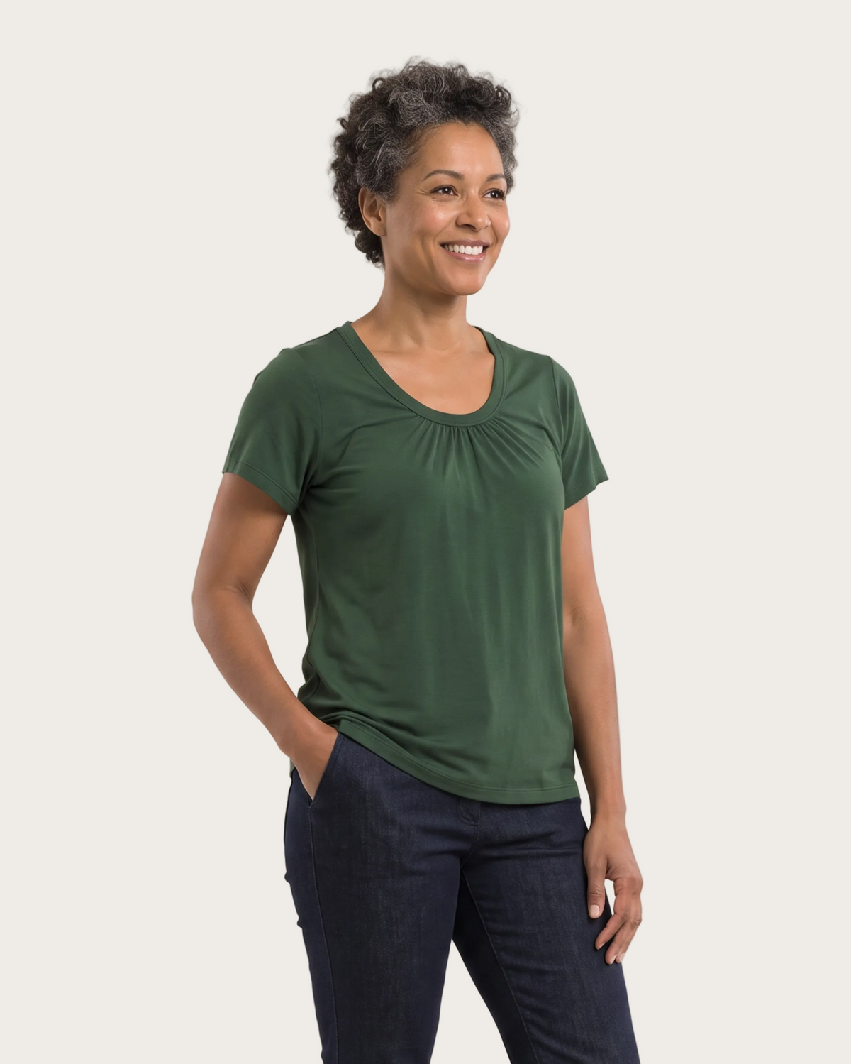 A woman modeling a dark green short-sleeve top with a gathered neckline and dark blue jeans, standing against a plain light gray studio background.