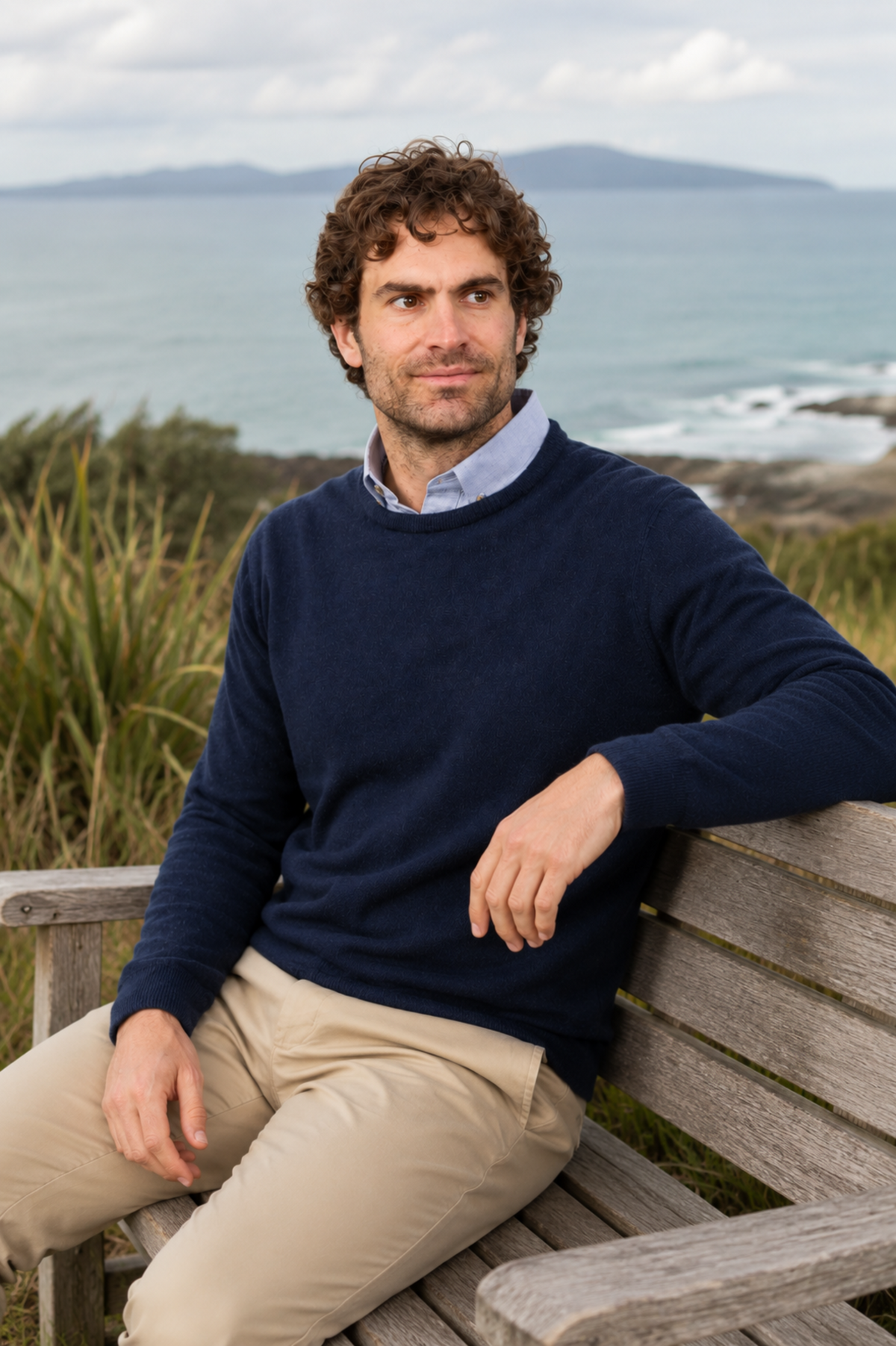 Man sitting on a wooden bench in a possum merino twilight jumper by the ocean with mountains in the background