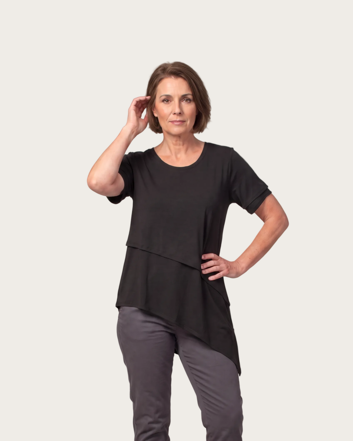 A woman modeling a black asymmetrical short-sleeve top with dark gray pants and brown leather shoes, standing against a solid taupe studio background.