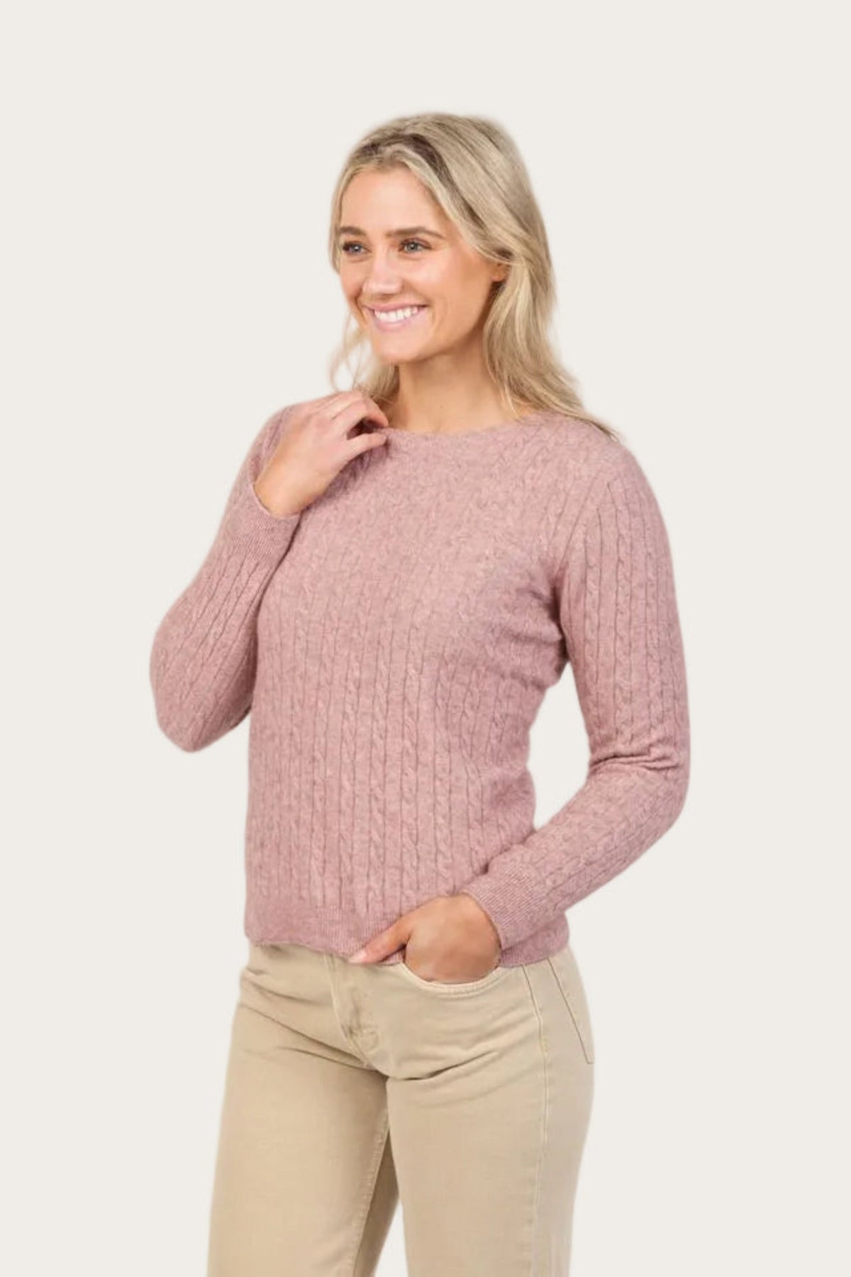 Woman wearing a pink possum merino cable knit sweater and beige pants on a white background