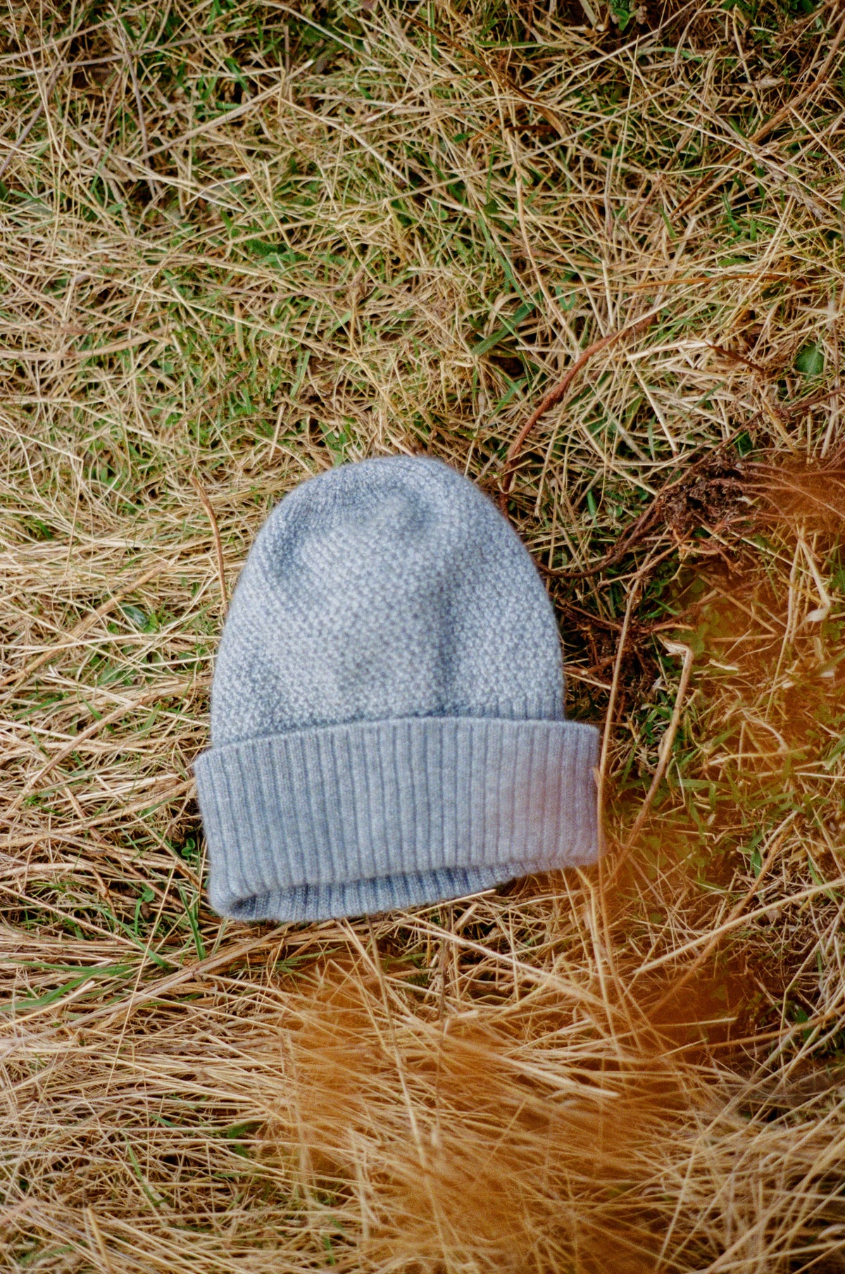 A light blue-gray knit beanie hat with a ribbed cuff, placed on a bed of dry brown grass and green grass.