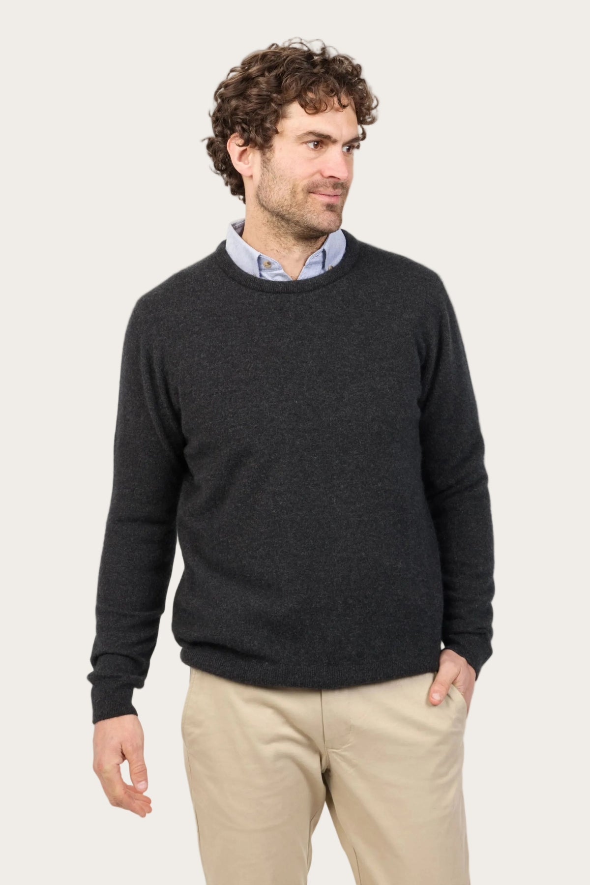 Man wearing a dark gray sweater over a collared shirt with beige pants on a white background